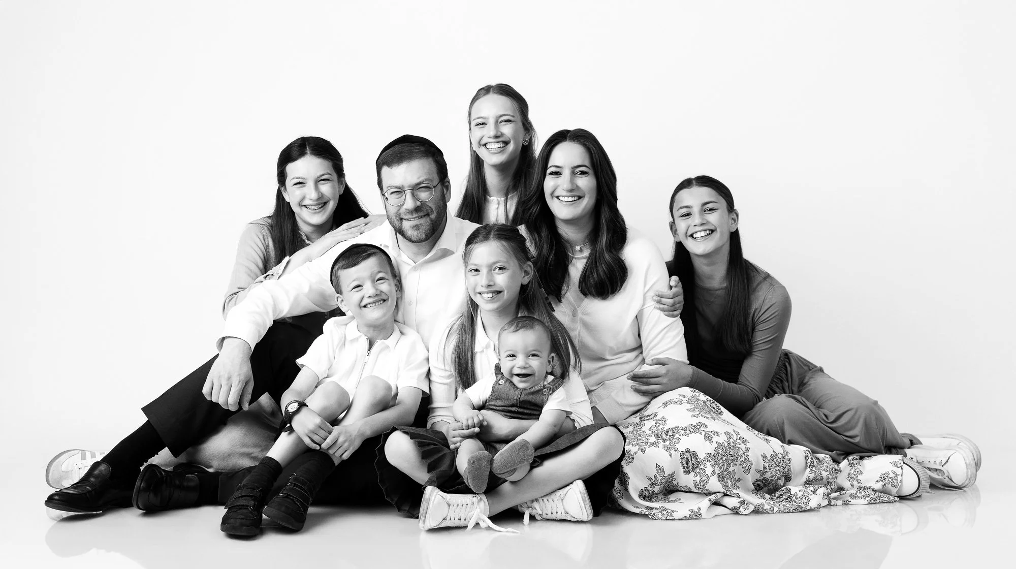 A black and white photo of a diverse family of ten, including adults and children, sitting and lying on the floor against a plain white background, smiling and looking at the camera.