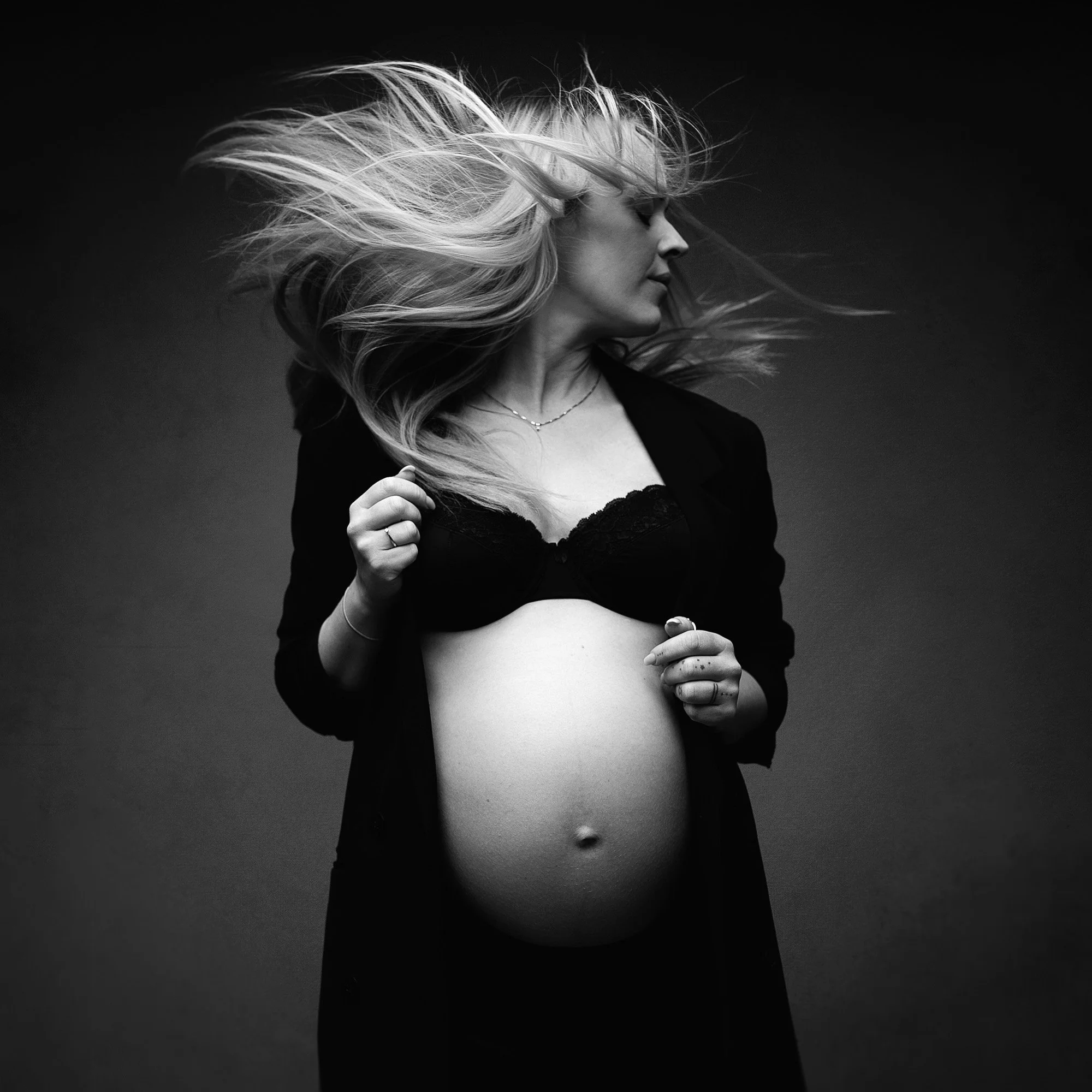 A pregnant woman with long hair flowing in the wind, wearing a black lingerie top and an open black jacket, holding her belly with both hands, in a black and white studio portrait.