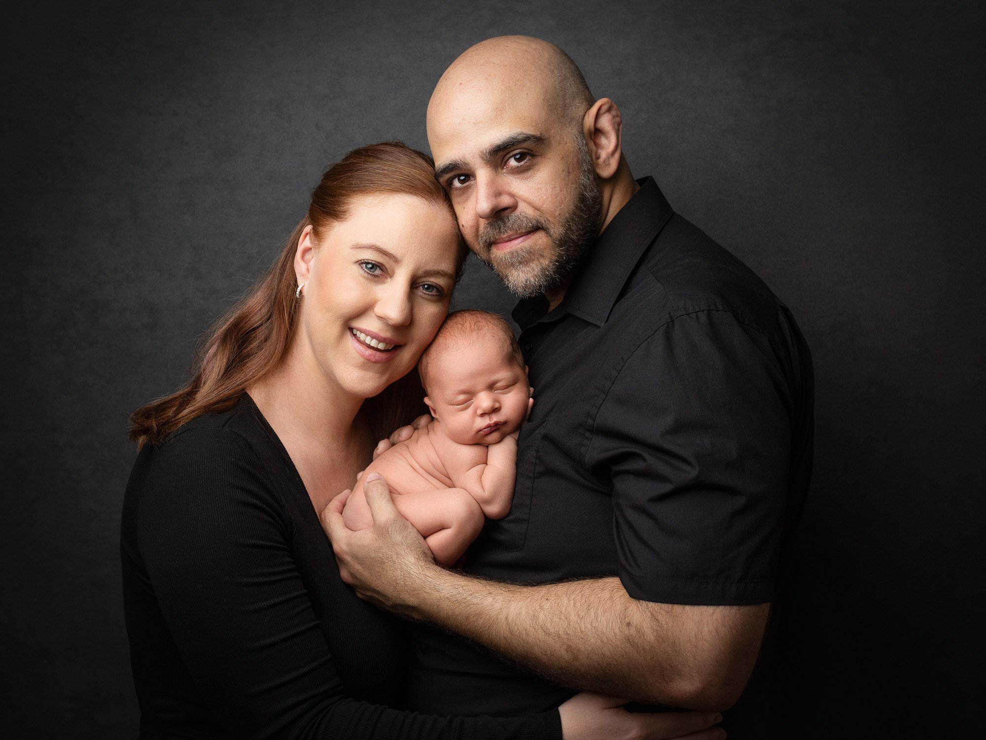 A family of three with a woman, a man, and a newborn baby, all dressed in black, posing against a dark background.