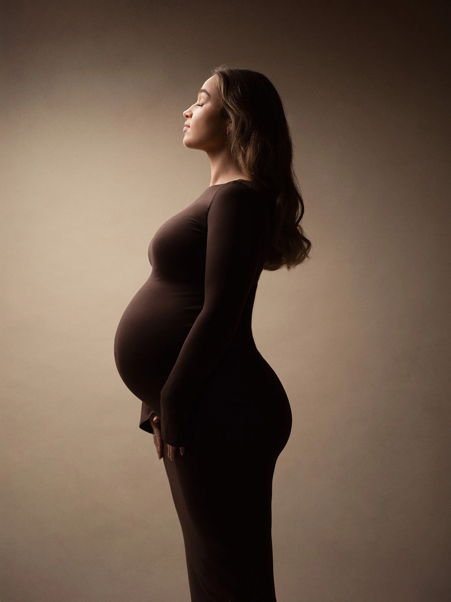 Profile of a pregnant woman with closed eyes, wearing a long, form-fitting brown dress, standing against a plain background.
