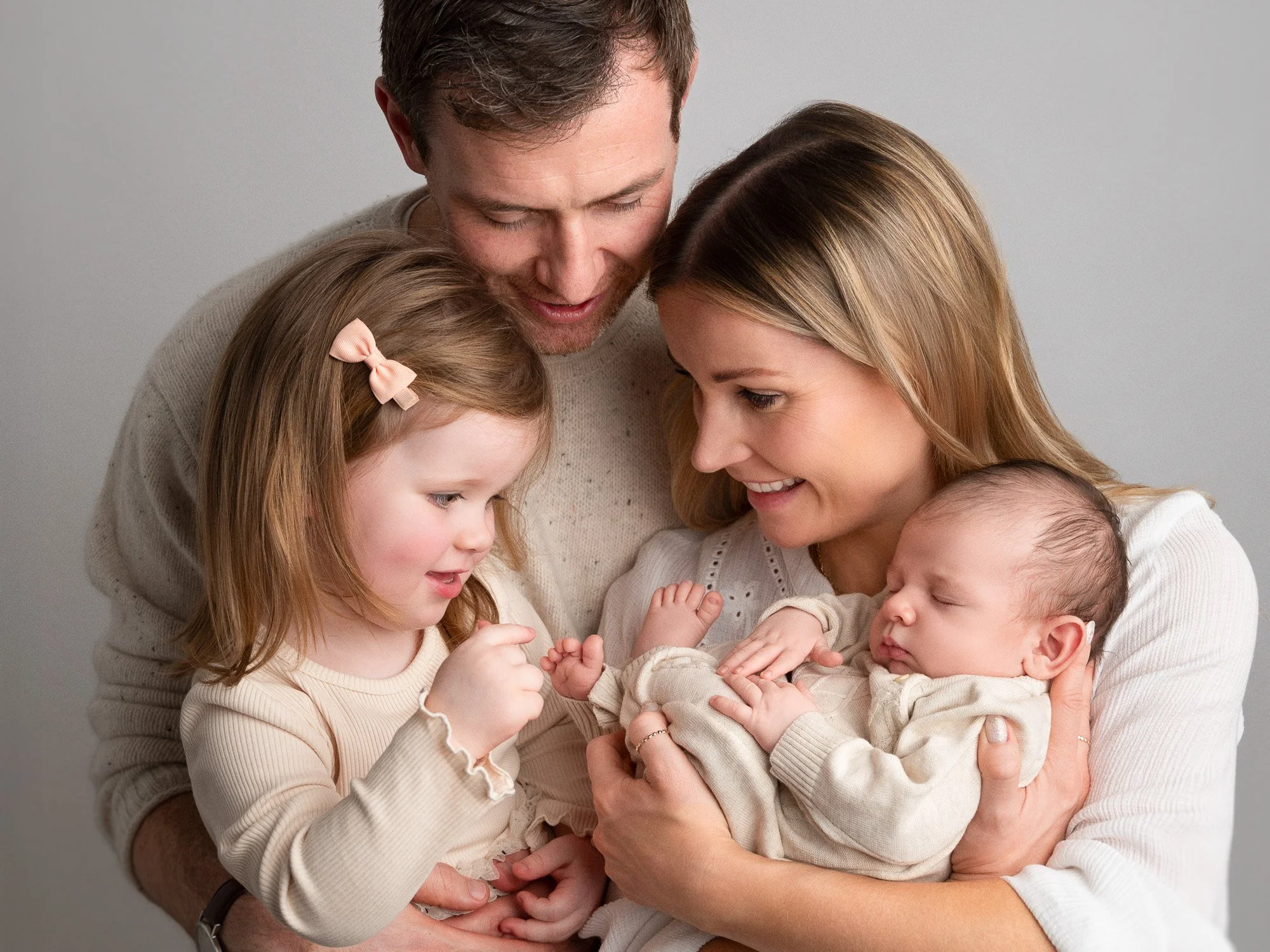 A family of four, including a man, woman, a young girl, and a sleeping baby, gathered closely together outdoors, smiling and showing affection.