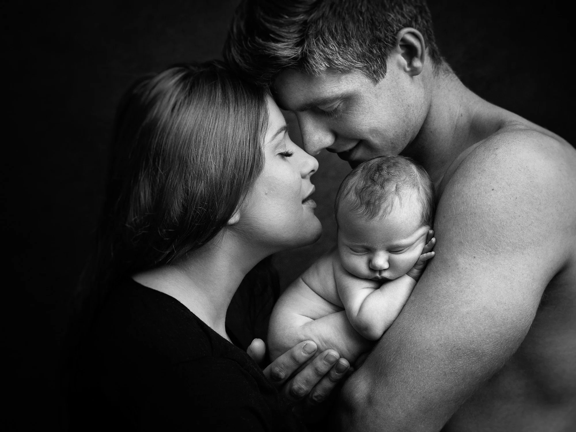 A black and white photo of a family, where a woman and a shirtless man gently hold a sleeping baby, their foreheads touching, creating an intimate moment.