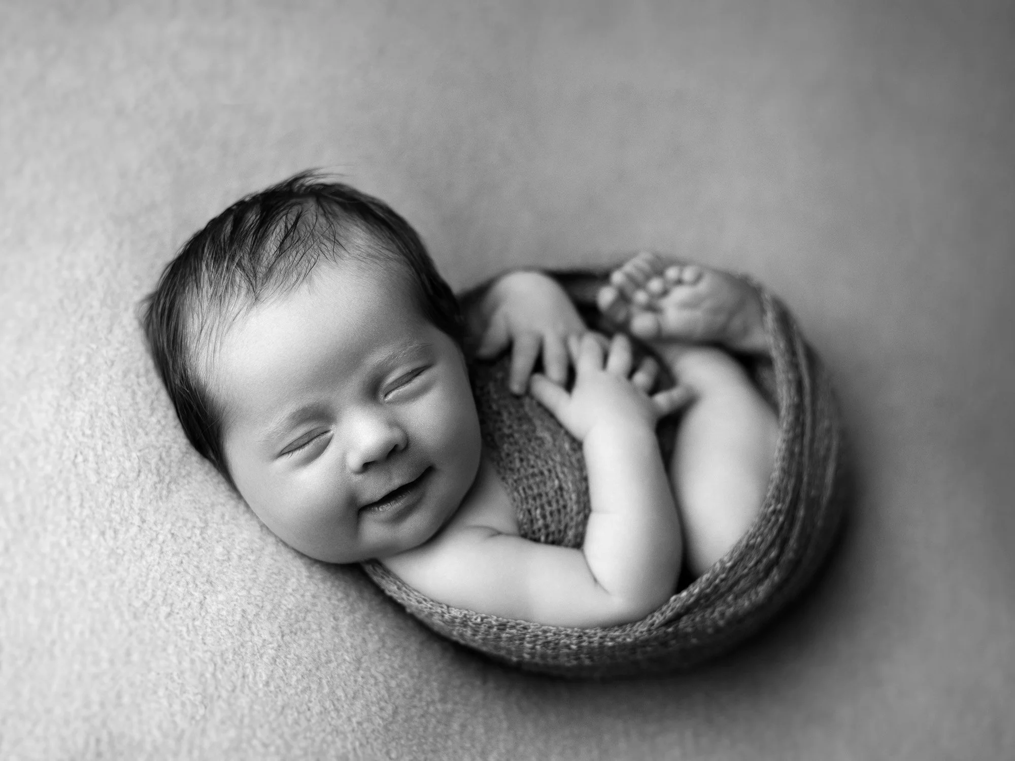 Black and white photo of a smiling baby with closed eyes, lying on soft textured surface, wrapped in a knitted blanket with feet visible.