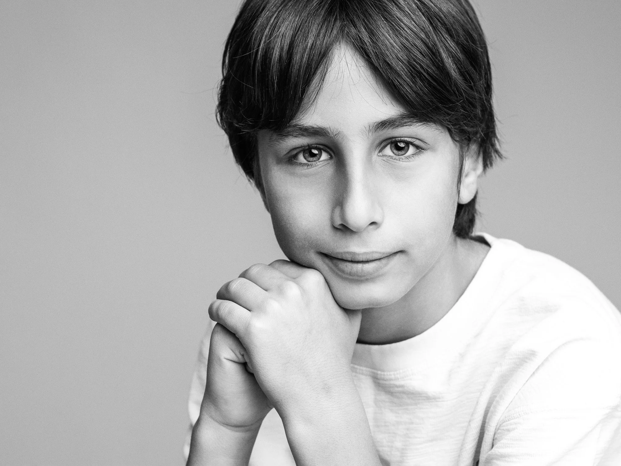 Black and white portrait of a young boy with medium-length hair, resting his chin on his clasped hands, looking directly at the camera with a subtle smile.