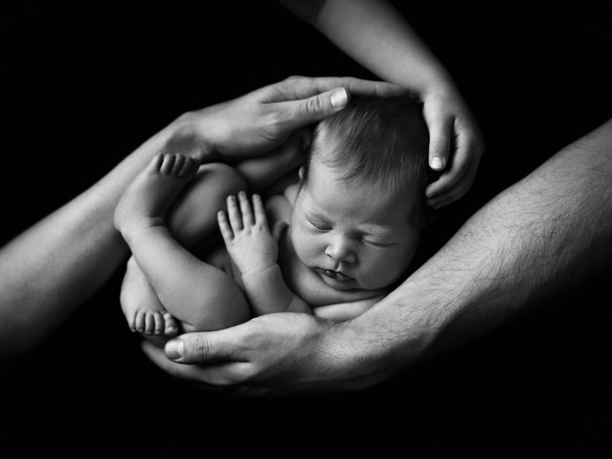 A black and white photo of a newborn cradled in multiple hands, with the baby's face relaxed and eyes closed.
