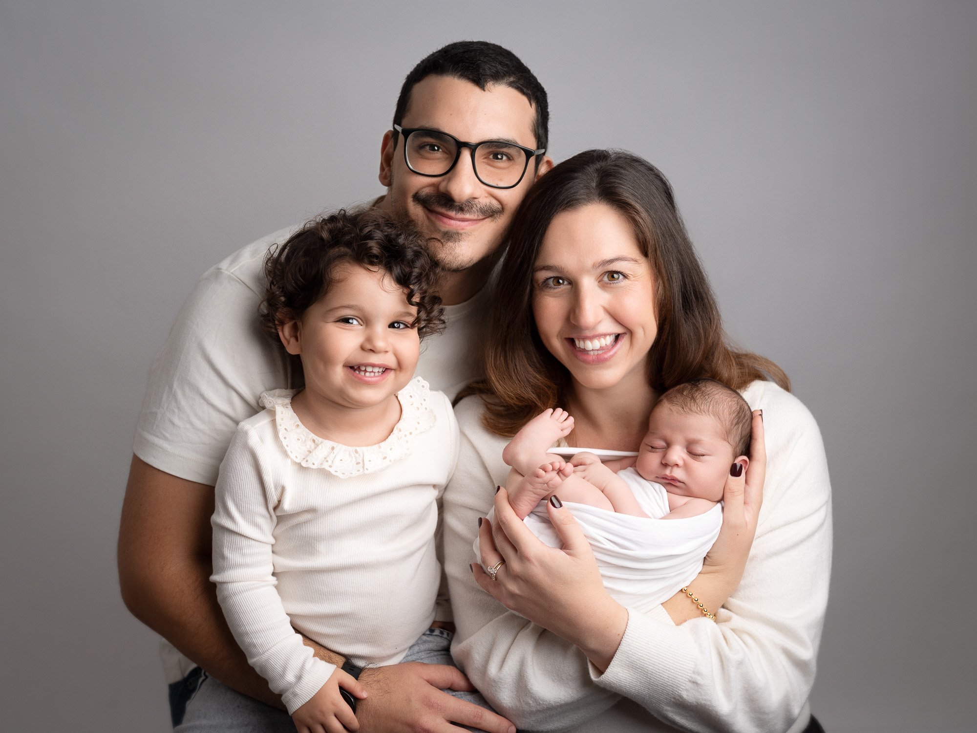 Happy family with father, mother, young daughter, and newborn baby smiling and posing together against a gray background.