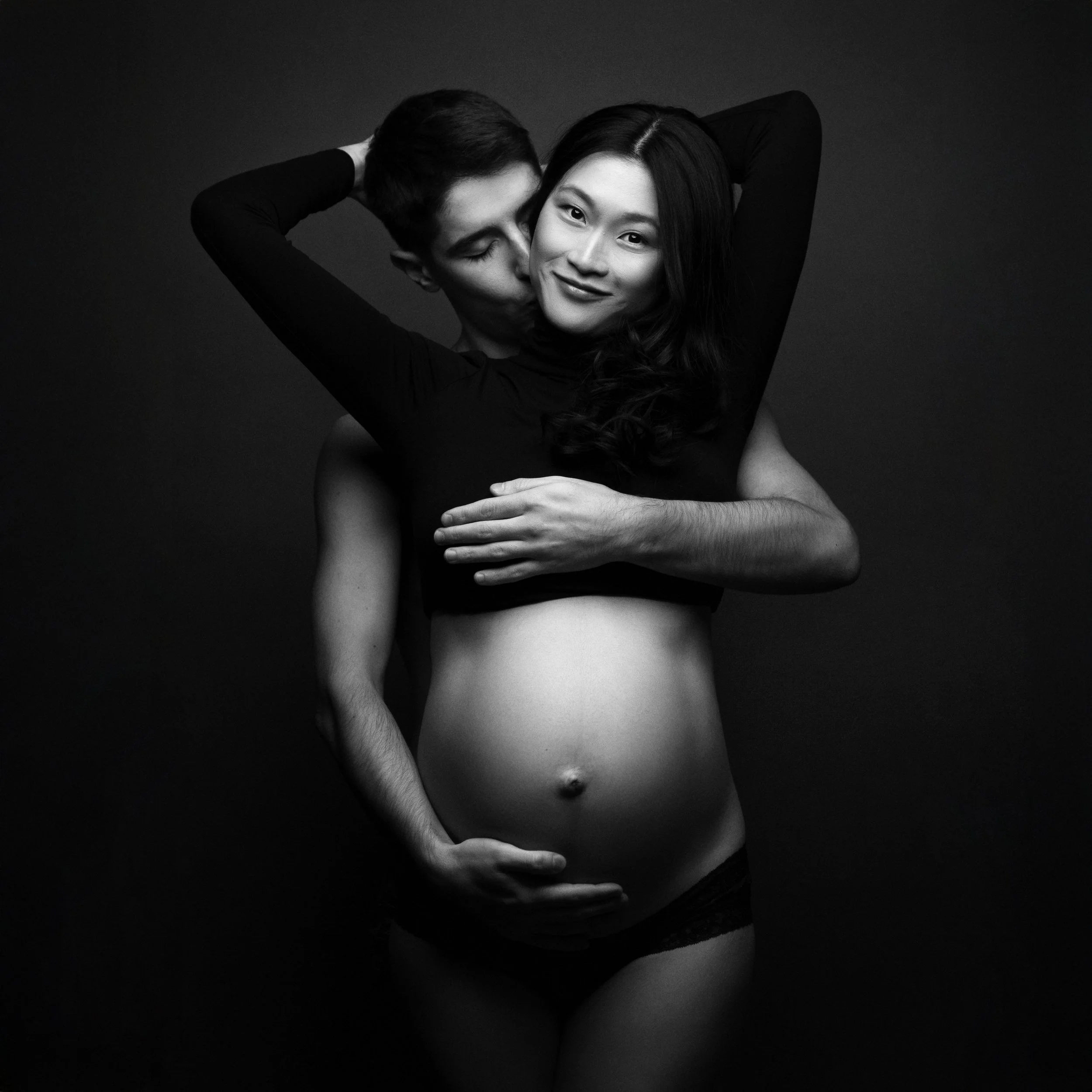 Black and white photo of a pregnant woman with long dark hair, smiling, with her hand on her belly, and her partner with short hair, kissing her cheek from behind, both holding her belly.