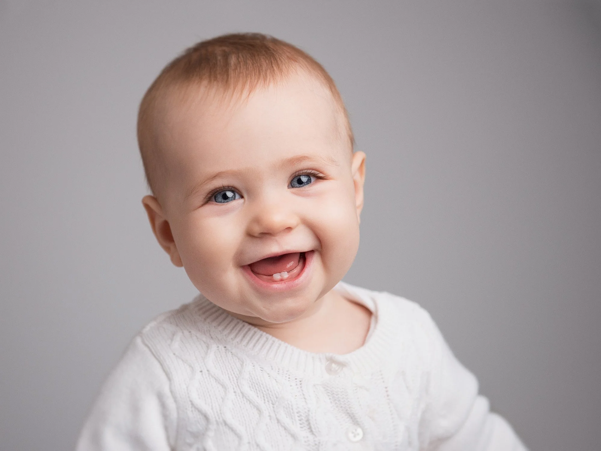 Close-up of a smiling toddler with blue eyes and red hair, wearing a white knit sweater, against a plain gray background.