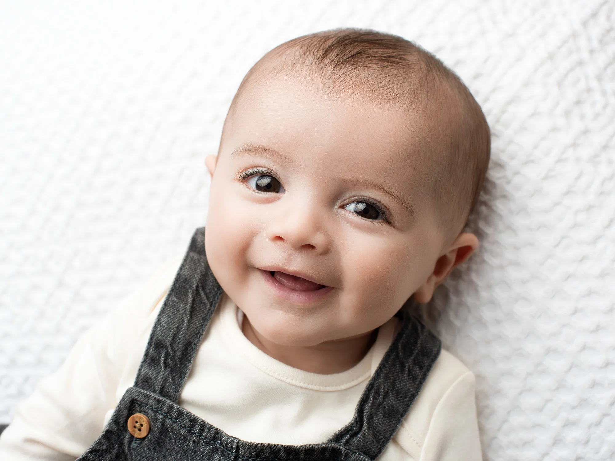 Smiling toddler with big brown eyes and short light brown hair lying on a white textured blanket, wearing a beige shirt and dark gray overalls.