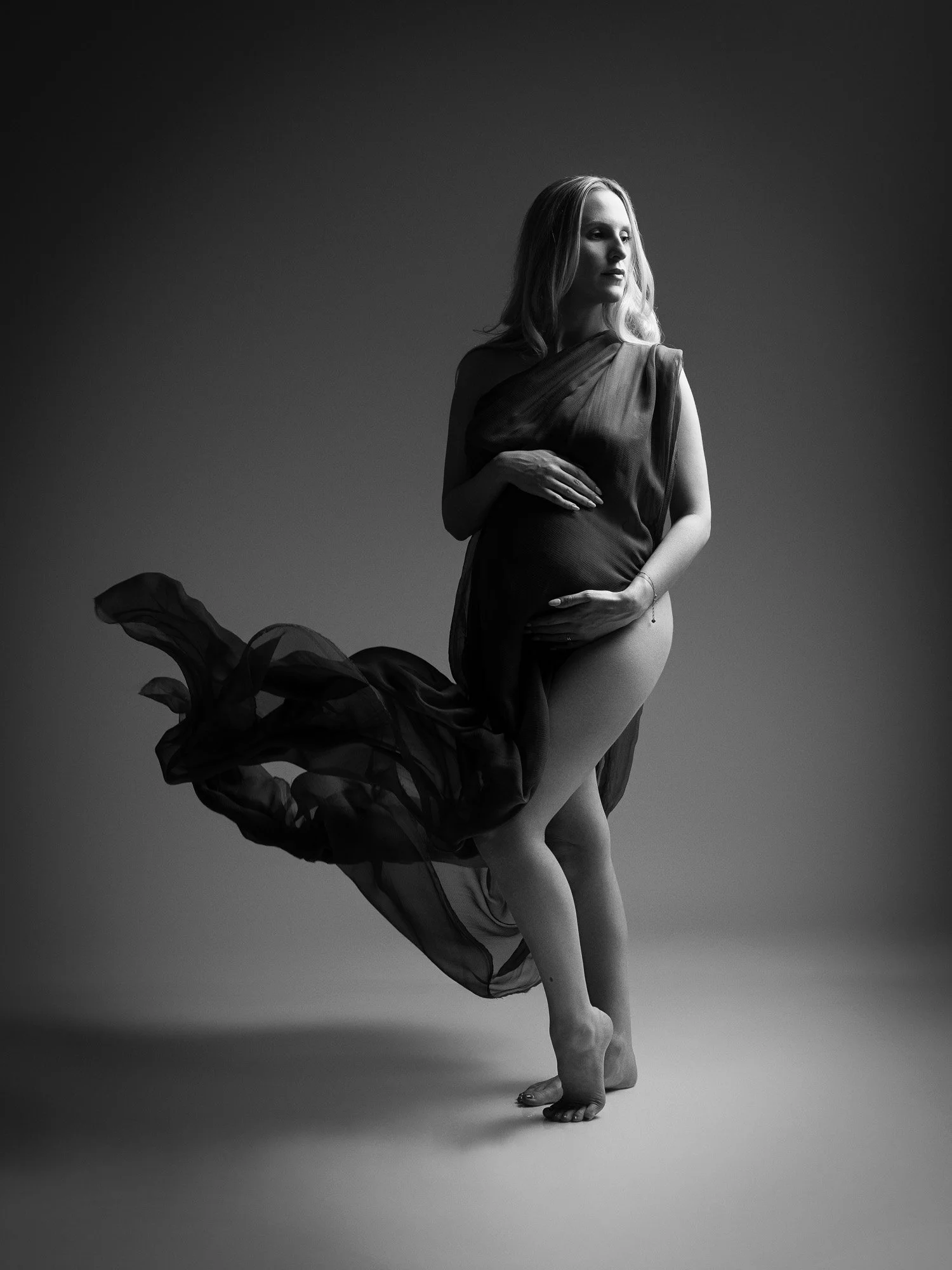 Black and white photo of a pregnant woman standing barefoot, with her left leg slightly bent and her right leg crossing in front of her. She has long, wavy hair and is wearing a sheer, flowing fabric draped over her shoulders and around her waist. He