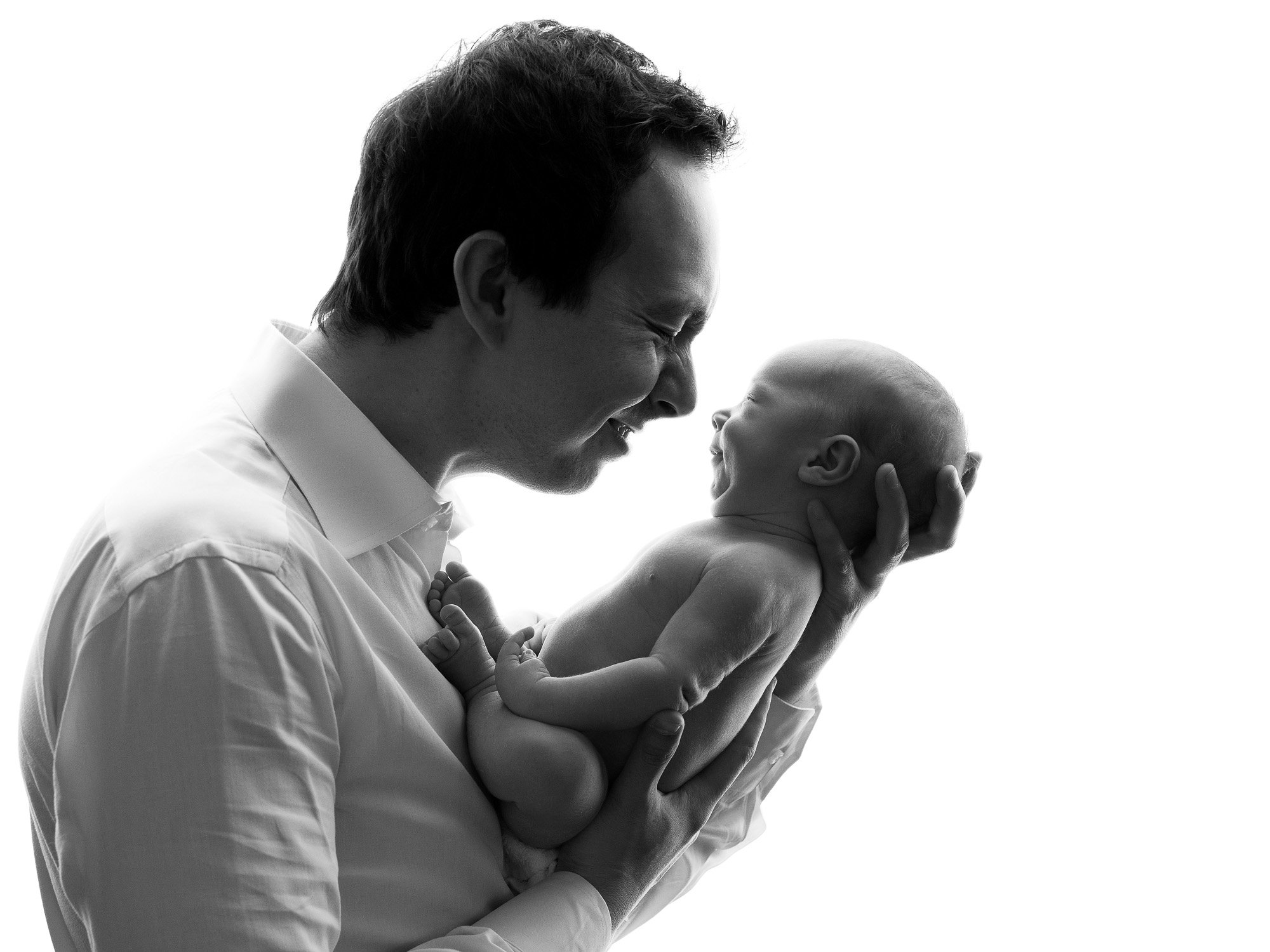 A man holding a newborn baby close to his face, smiling with their foreheads touching in silhouette.