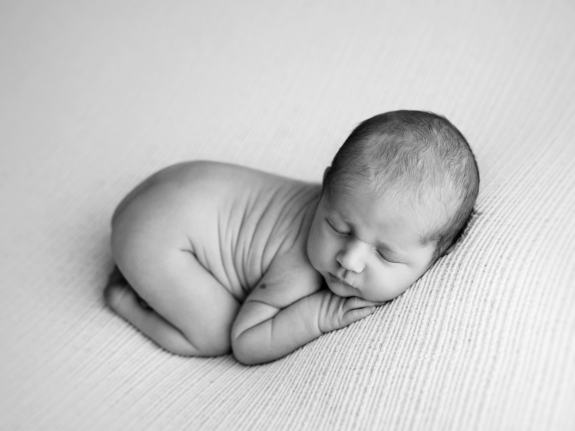 Black and white photo of a newborn baby lying on a soft surface with head resting on hands, eyes closed, and body curled up.