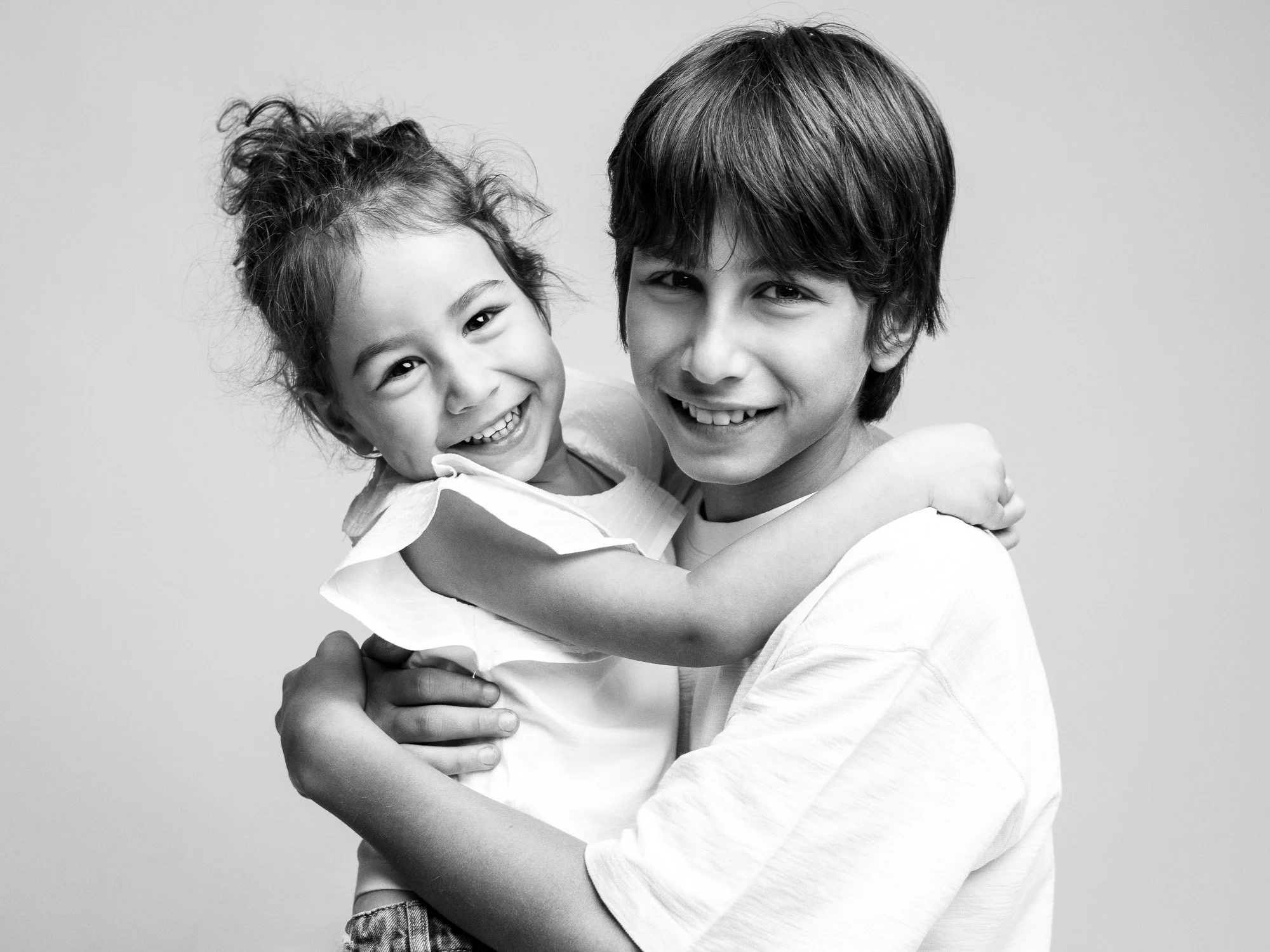A young boy with medium-length dark hair smiling while holding a young girl with curly hair in a ponytail, smiling widely and hugging him, against a plain background.