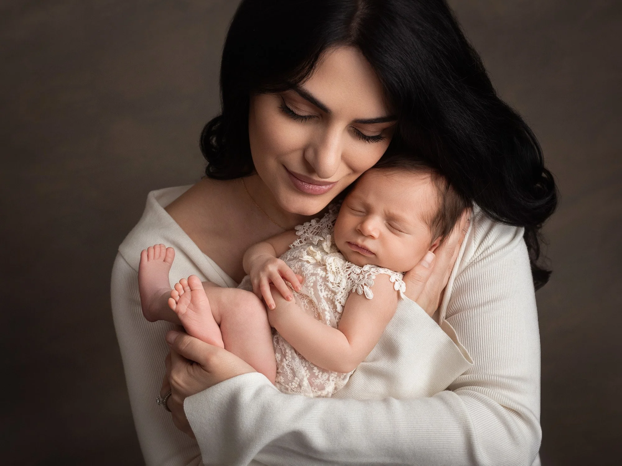 A woman with dark hair and a light-colored top holding a sleeping baby with dark hair, dressed in a lace outfit, close to her face against a neutral background.