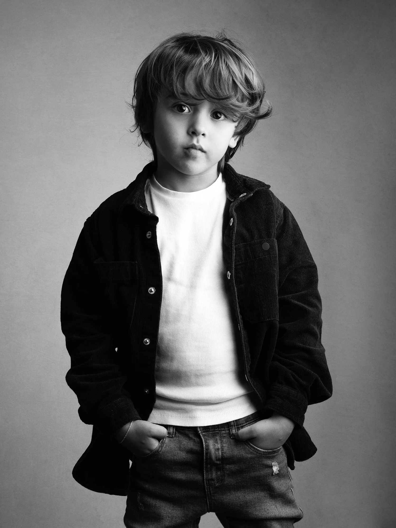 A young boy with wavy hair wearing a white t-shirt, dark jacket, and jeans, standing with hands in pockets against a plain background in black and white.