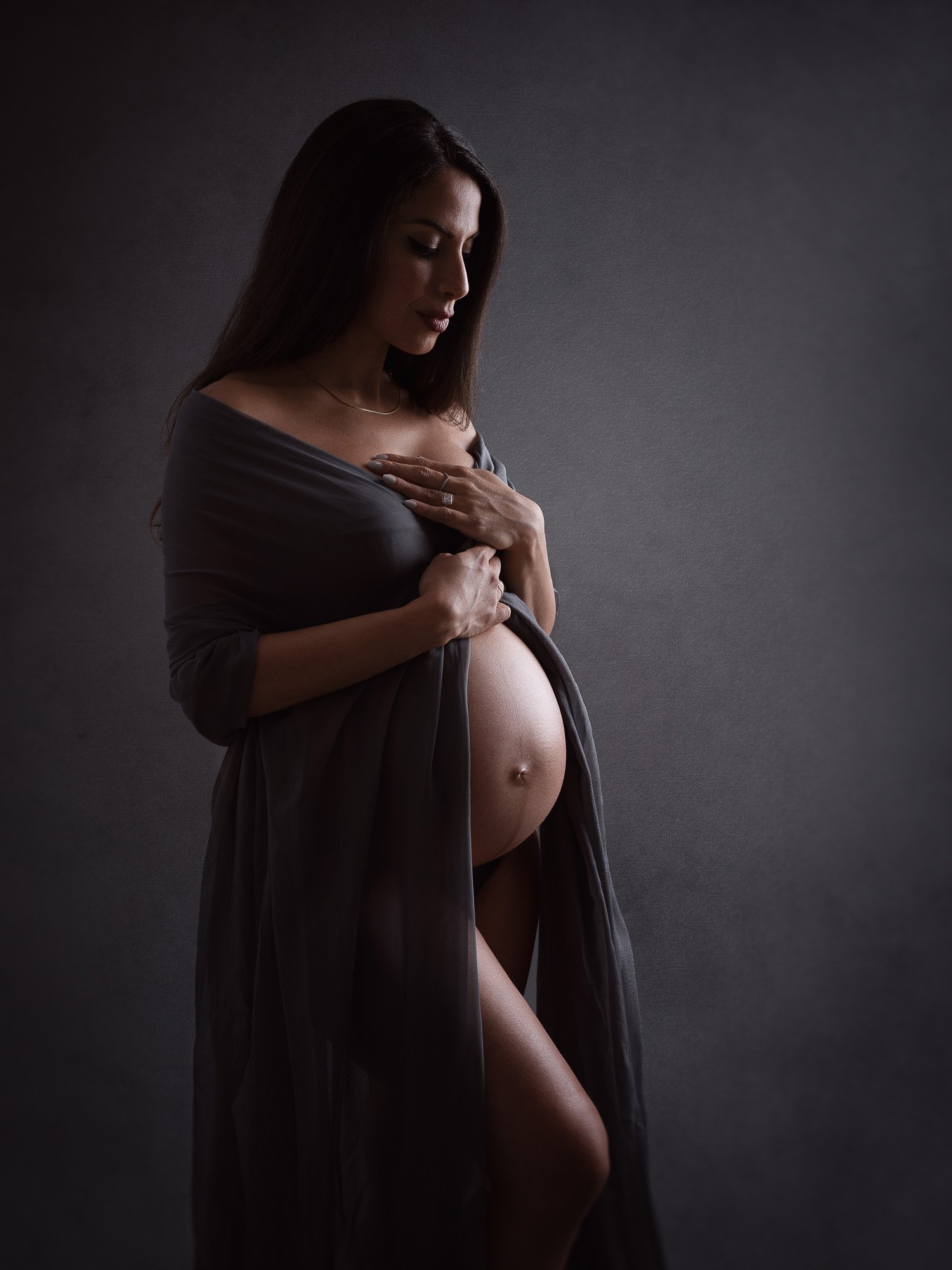 A pregnant woman standing against a dark gray background, wearing a semi-transparent gray dress that reveals her pregnant belly, with her hands resting on her chest and belly, looking down thoughtfully.