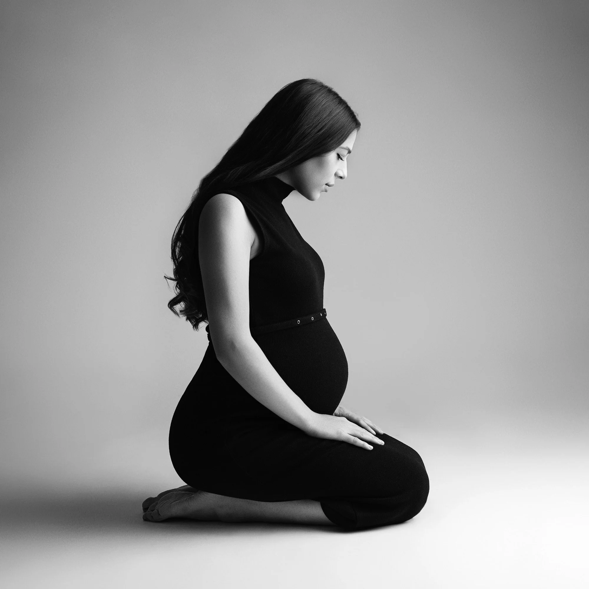 A pregnant woman kneeling on the ground with her eyes closed and hands resting on her legs, in a black dress and long hair, in black-and-white.
