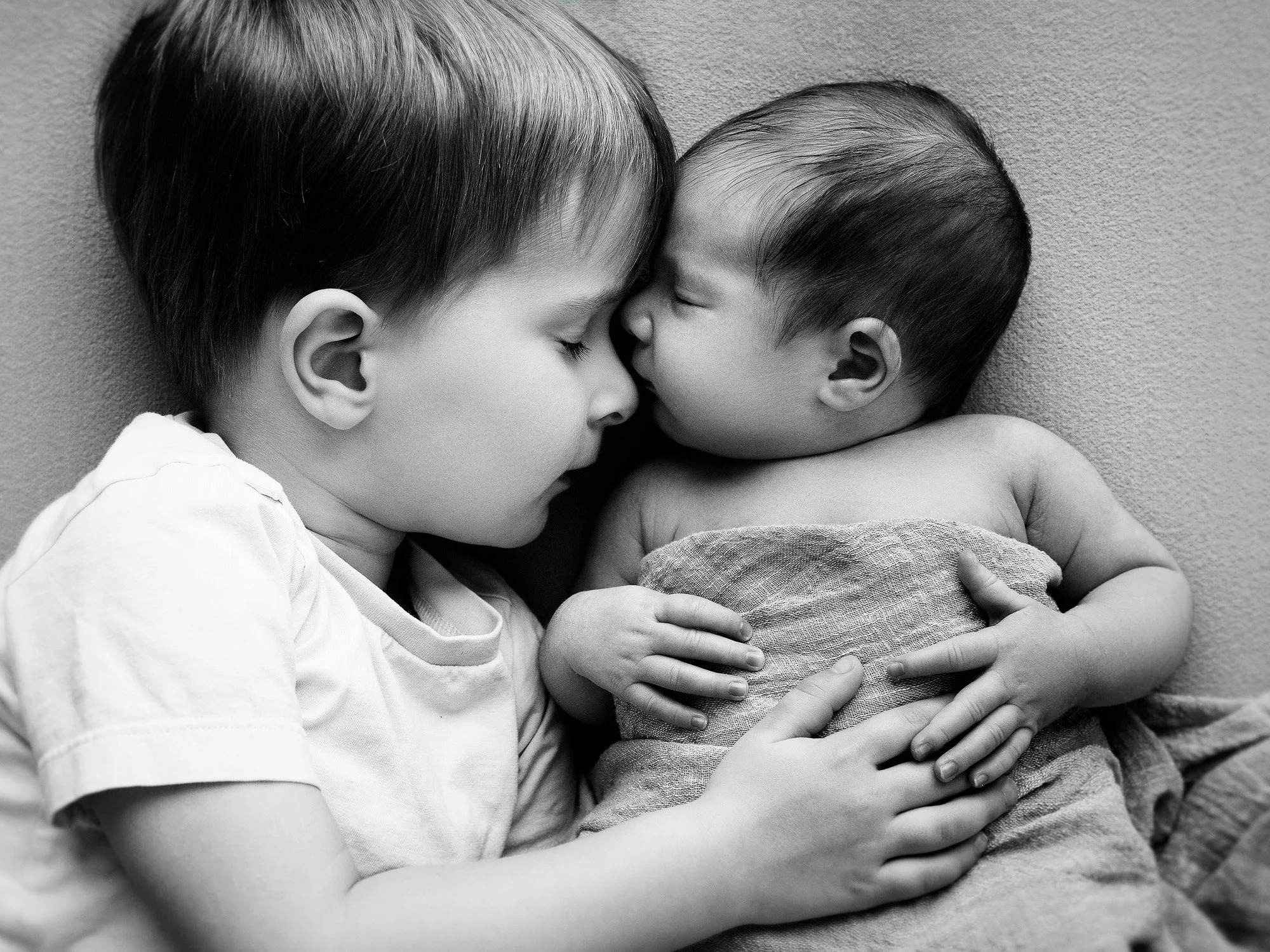 A black and white photo of a young boy and a baby lying close together face to face, touching noses, with the older child gently holding the baby.