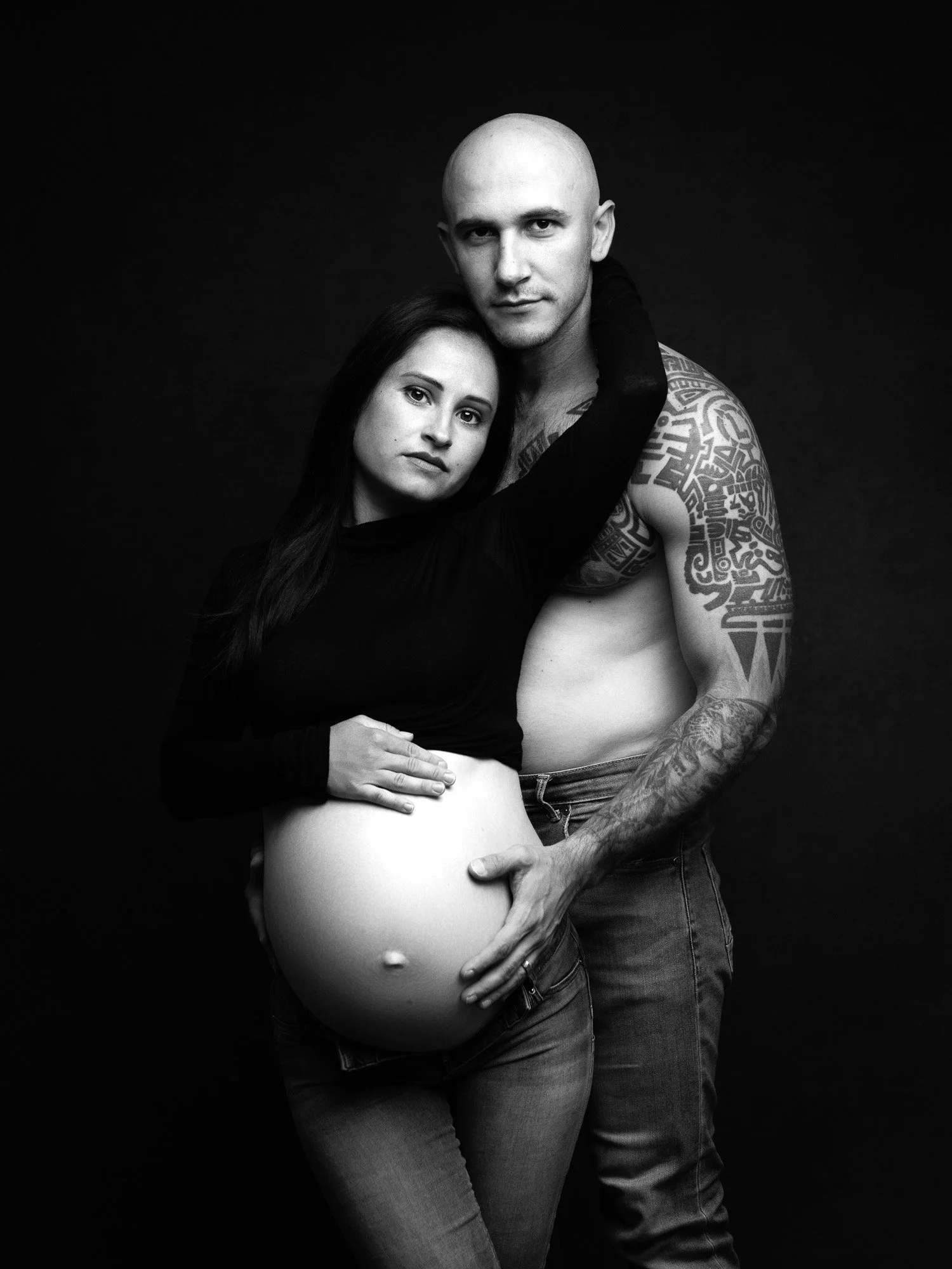 Black and white photo of a pregnant woman with long dark hair and a man with a shaved head. The woman is wearing a black long-sleeve shirt and the man is shirtless, exposing tattoos on his arm and chest. They are embracing, with the woman's arm aroun