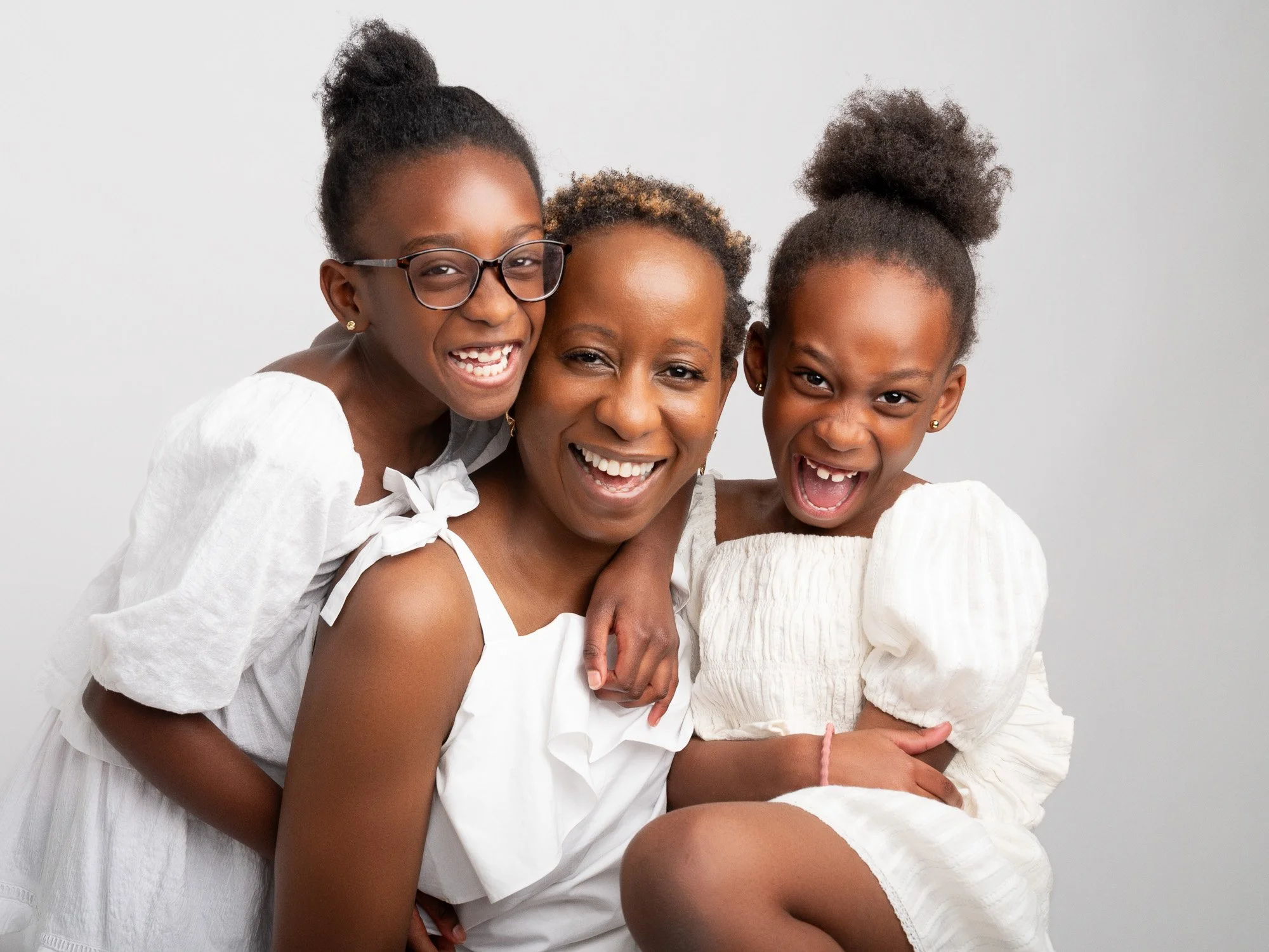Mother smiling with two young daughters, all wearing white dresses, posing together against a plain background.