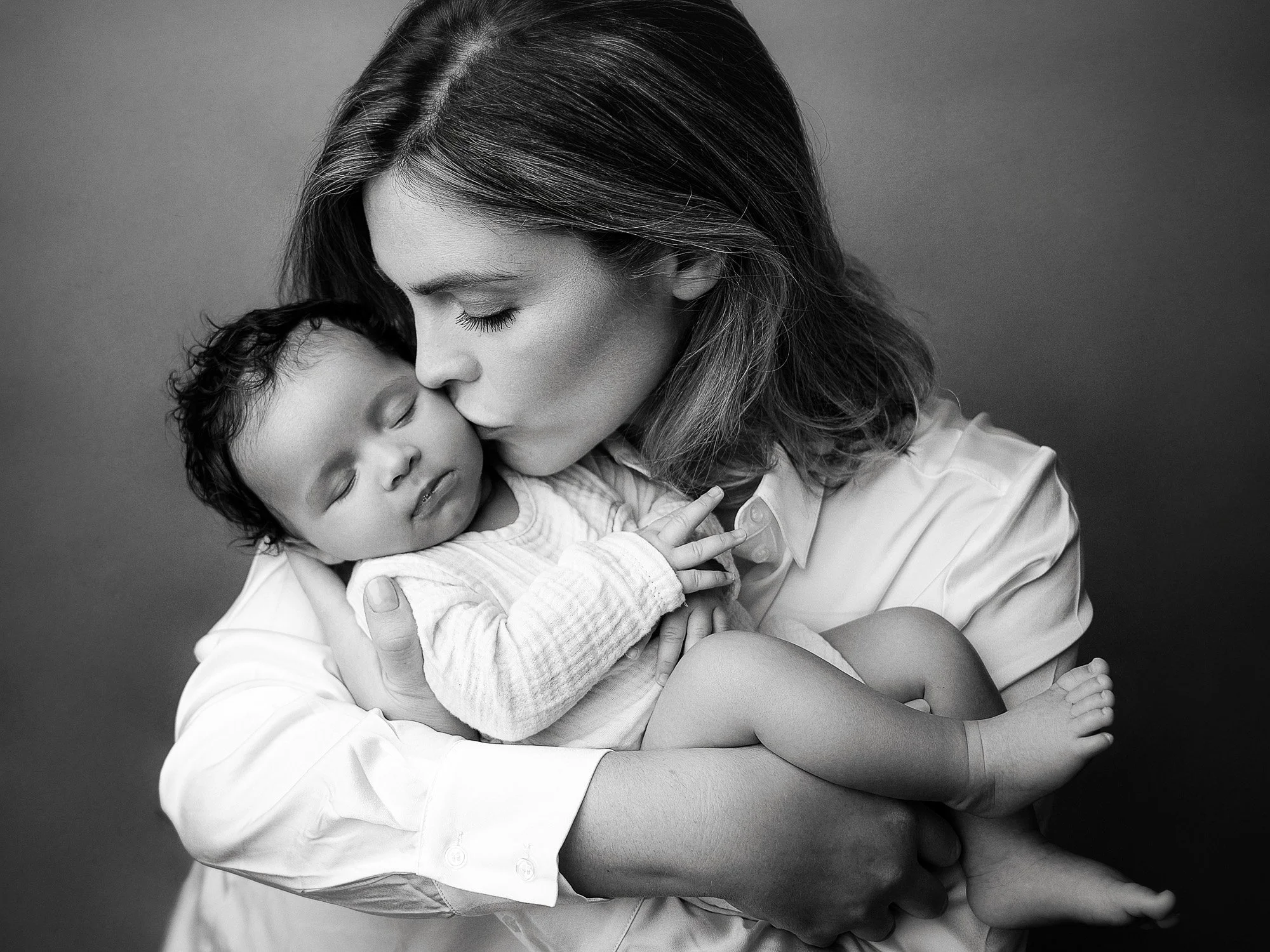 Black and white photo of a woman gently holding a sleeping young child, kissing their forehead, both with their eyes closed.