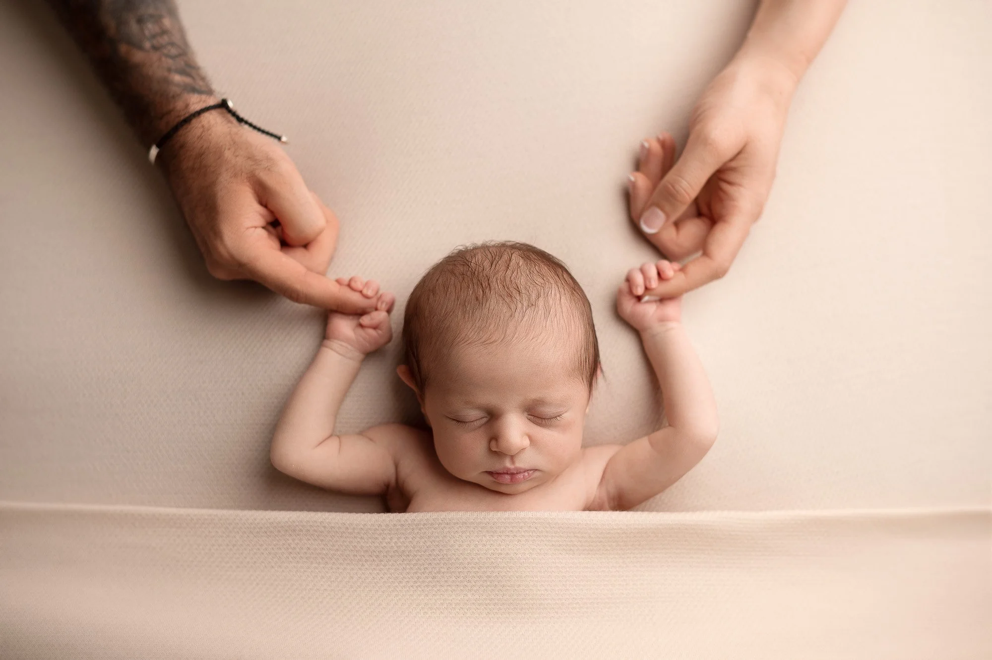 A newborn baby sleeping on a beige surface, with hands gently holding the baby's hands, one hand on each side.