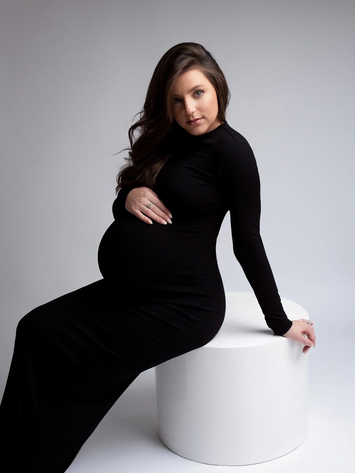 A pregnant woman in a black long-sleeve dress sitting on a white circular platform against a plain gray background.