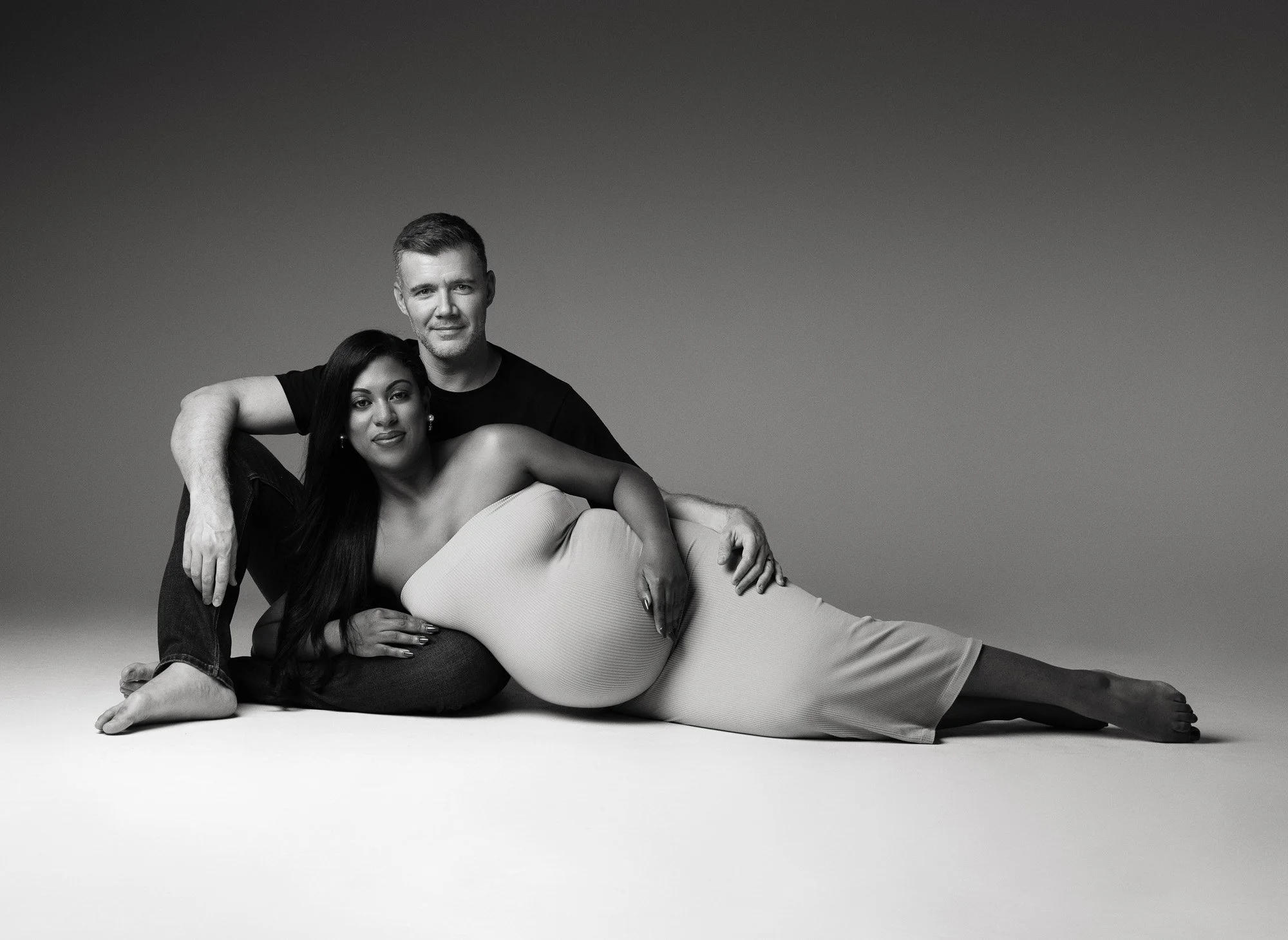 Black-and-white photograph of a man and a pregnant woman lying on the floor, with the woman resting her head on the man's lap and both looking at the camera.