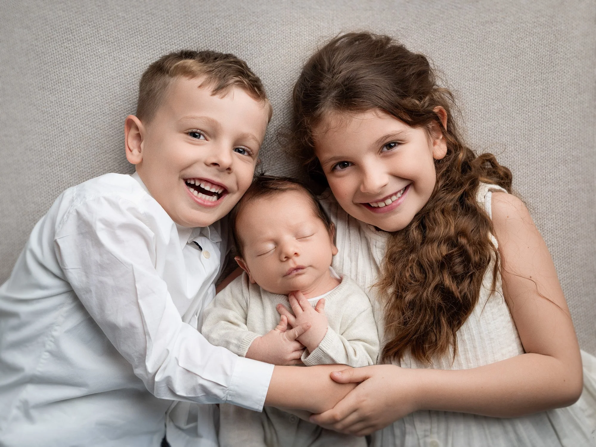 Three children, two girls and a boy, smiling and holding a newborn baby on a beige couch.