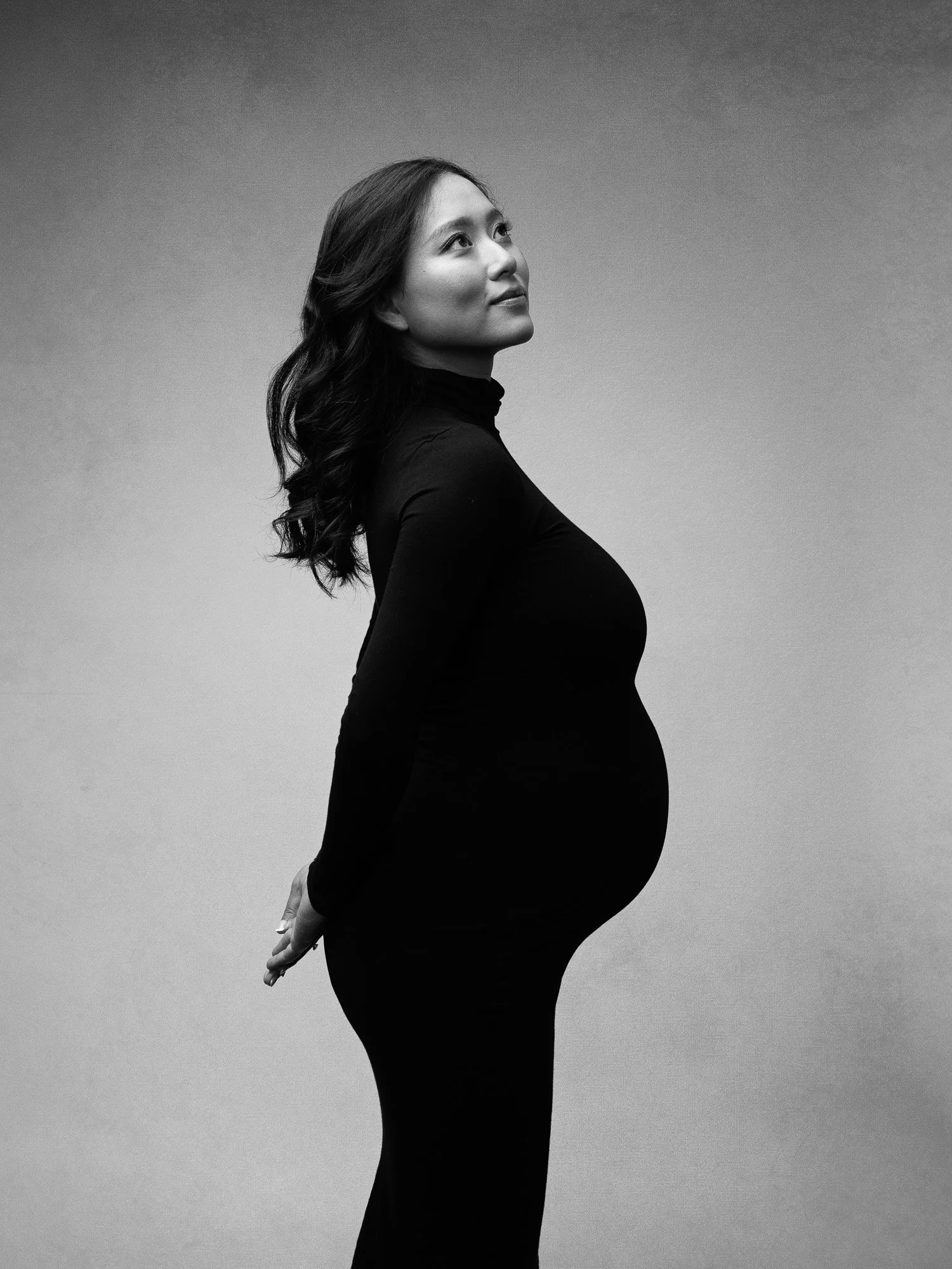 Black and white portrait of a pregnant woman in a black turtleneck, standing in profile with her hands behind her back, looking upward with a serene expression.