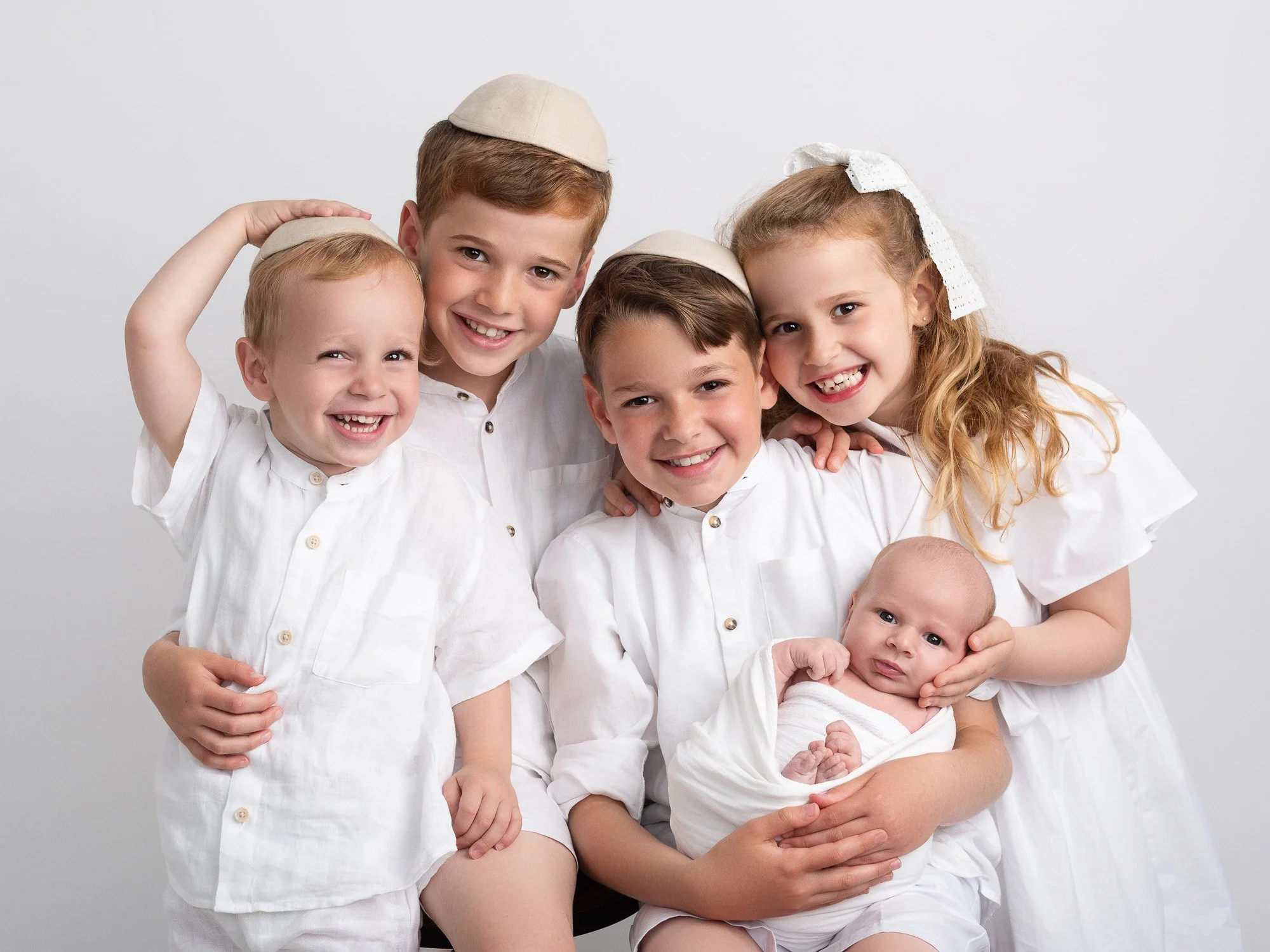 A group of six smiling children, four boys and two girls, dressed in white, with some wearing beige caps, posing together against a plain gray background.