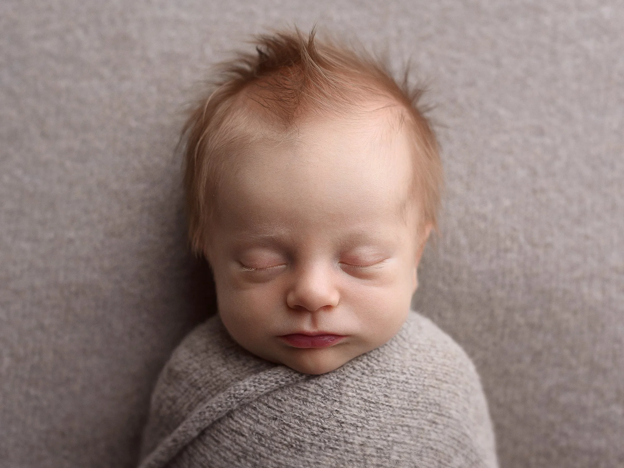 A close-up of a sleeping infant with light-colored hair, wrapped in a gray blanket, resting on a neutral-colored surface.