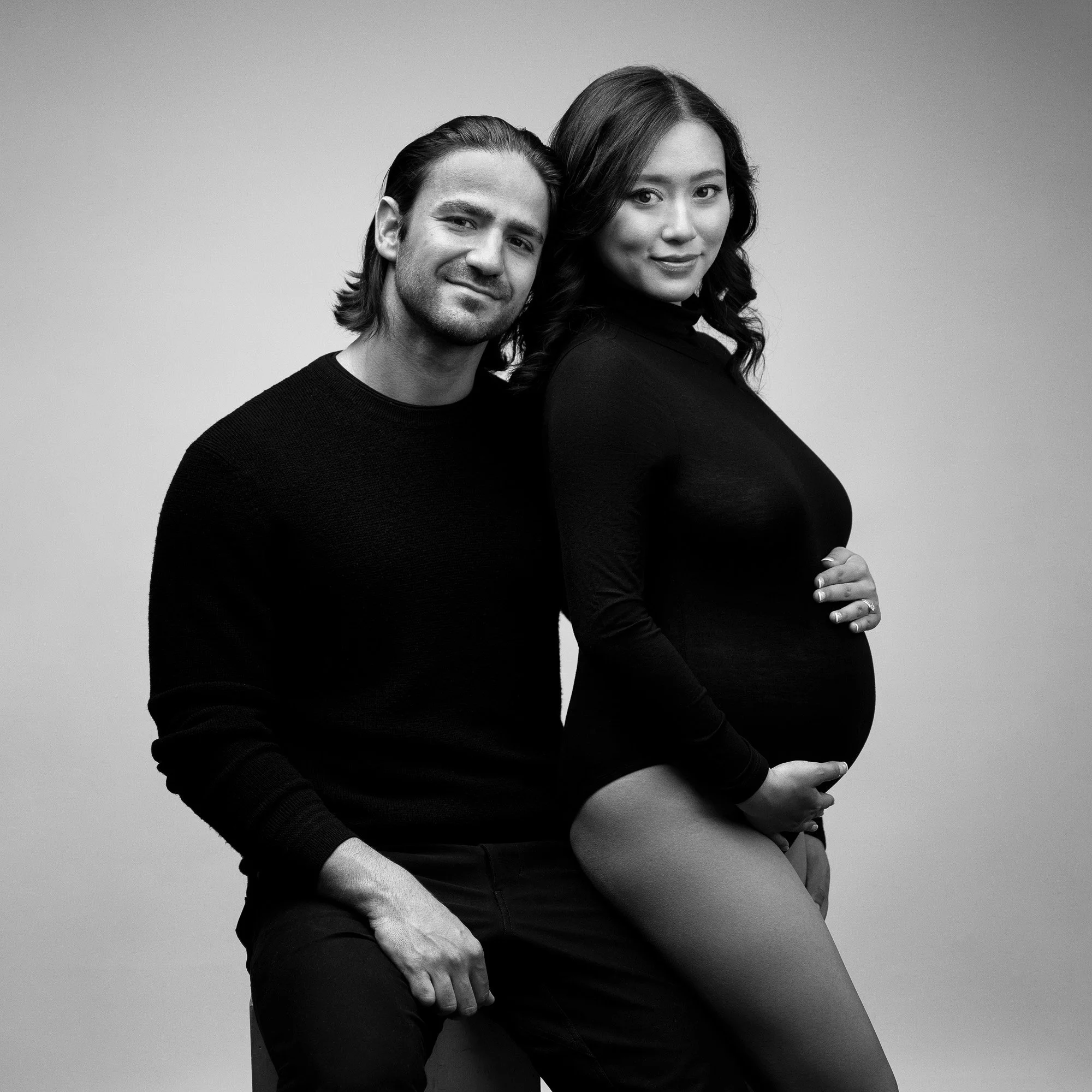 Black and white portrait of a pregnant woman sitting on a stool with a man standing beside her, both looking at the camera.