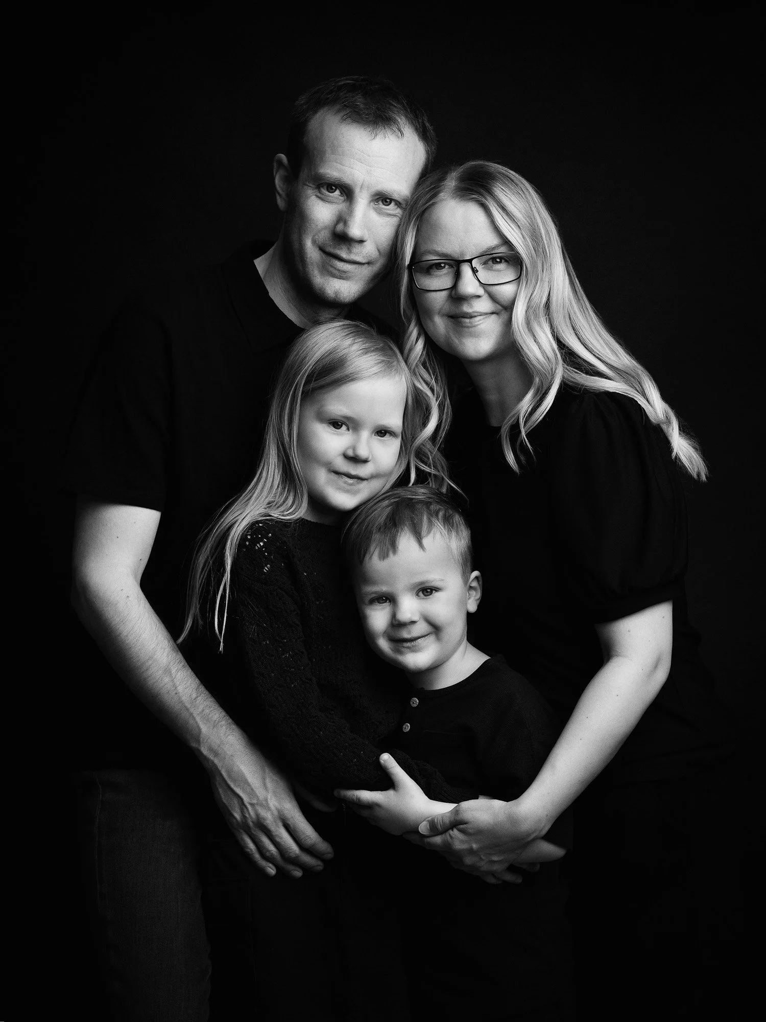 Black and white portrait of a family of four, including a man, a woman with glasses, a girl, and a boy, standing closely together and smiling.