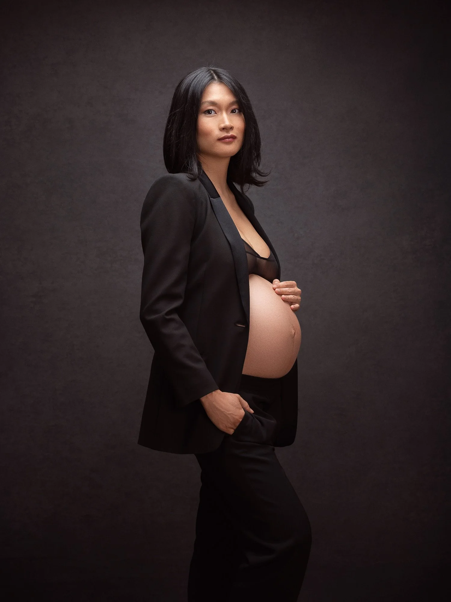 A pregnant woman with shoulder-length black hair wearing a black blazer and black pants, holding her belly with one hand, against a dark background.