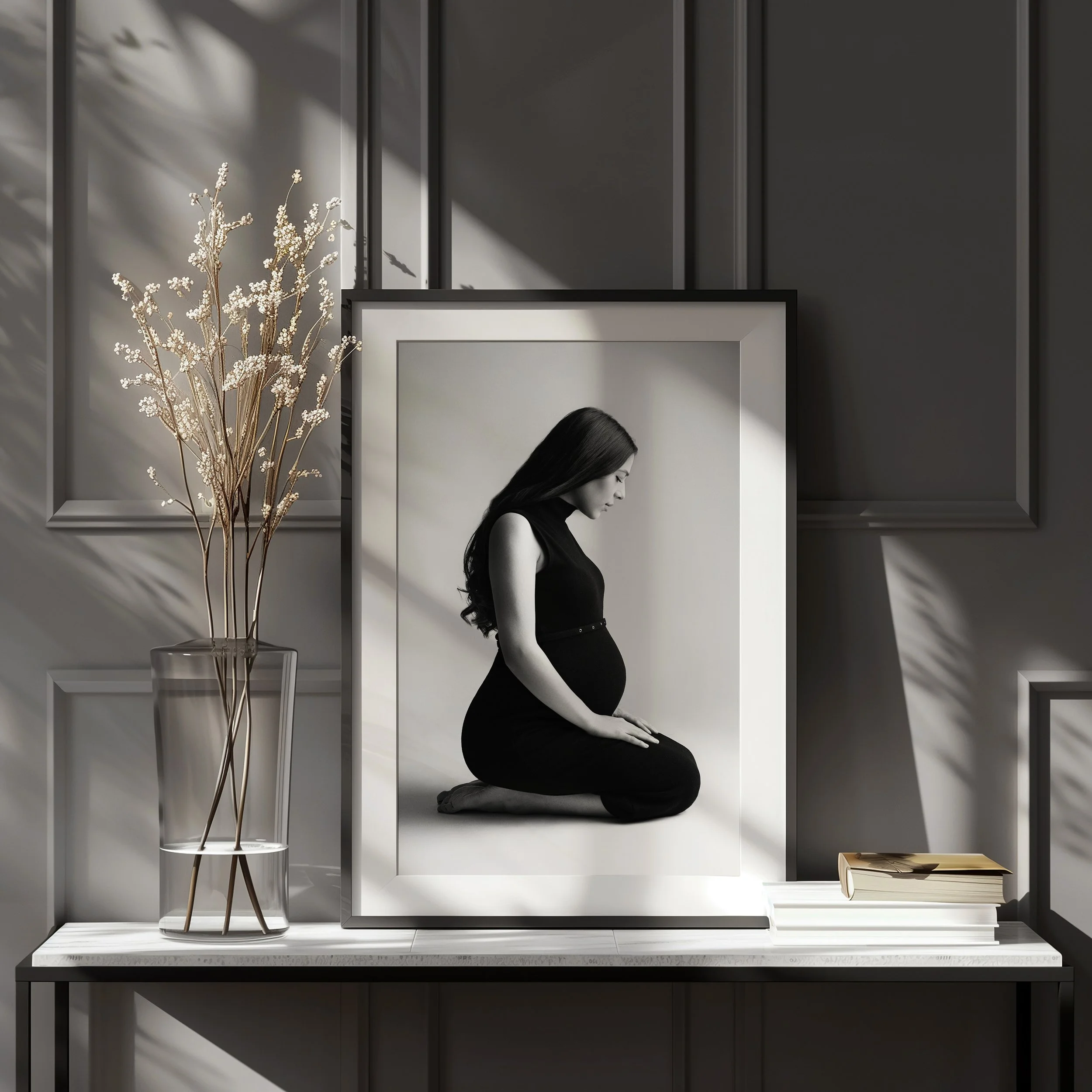 A black and white photograph of a pregnant woman kneeling with her head bowed and eyes closed inside a black frame, placed on a white shelf with a clear glass vase holding dried flowers to the left and a stack of books to the right against a gray paneled wall.