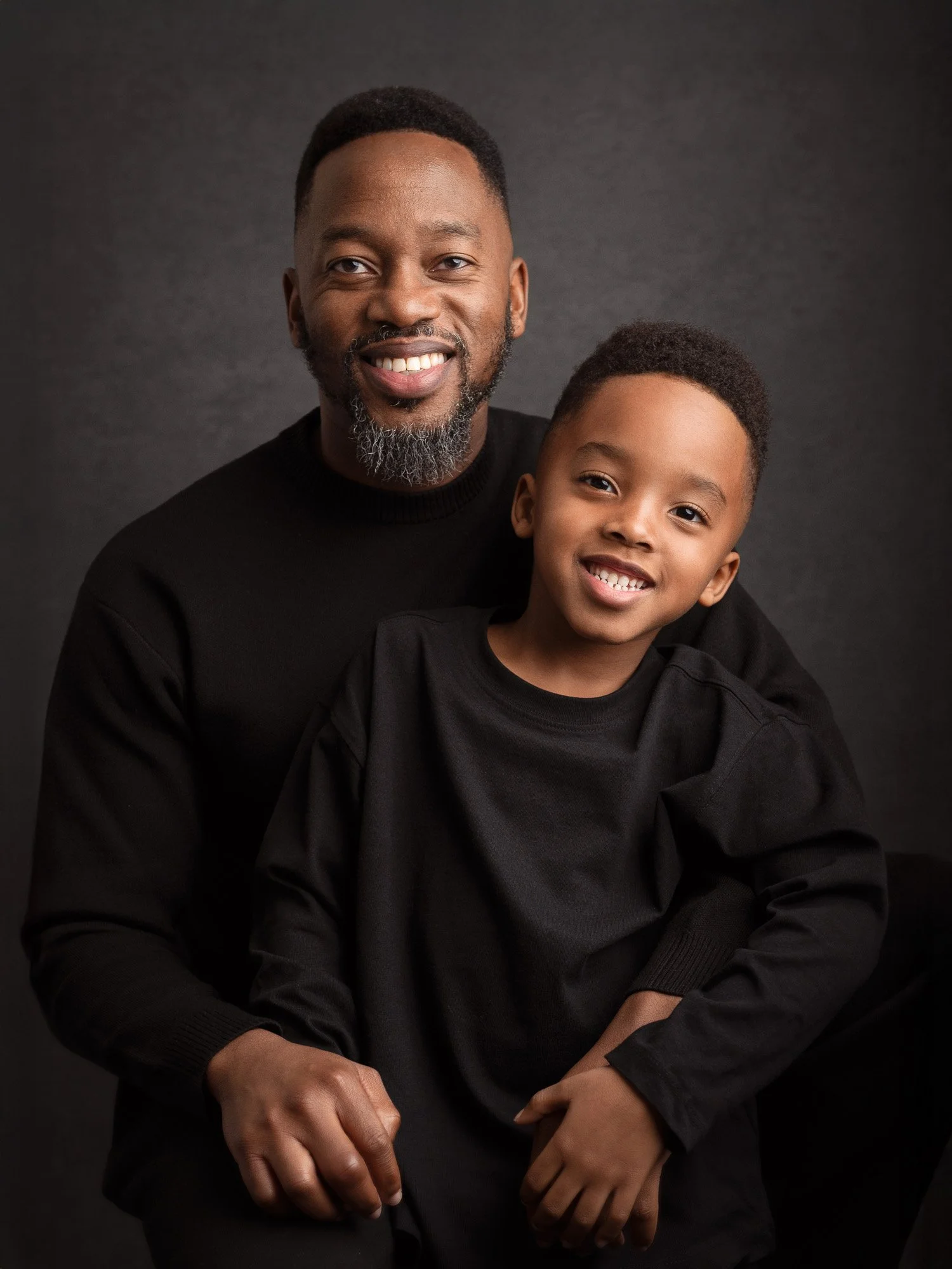A smiling African American man with a beard and a young boy, both wearing black shirts, sitting against a dark background.