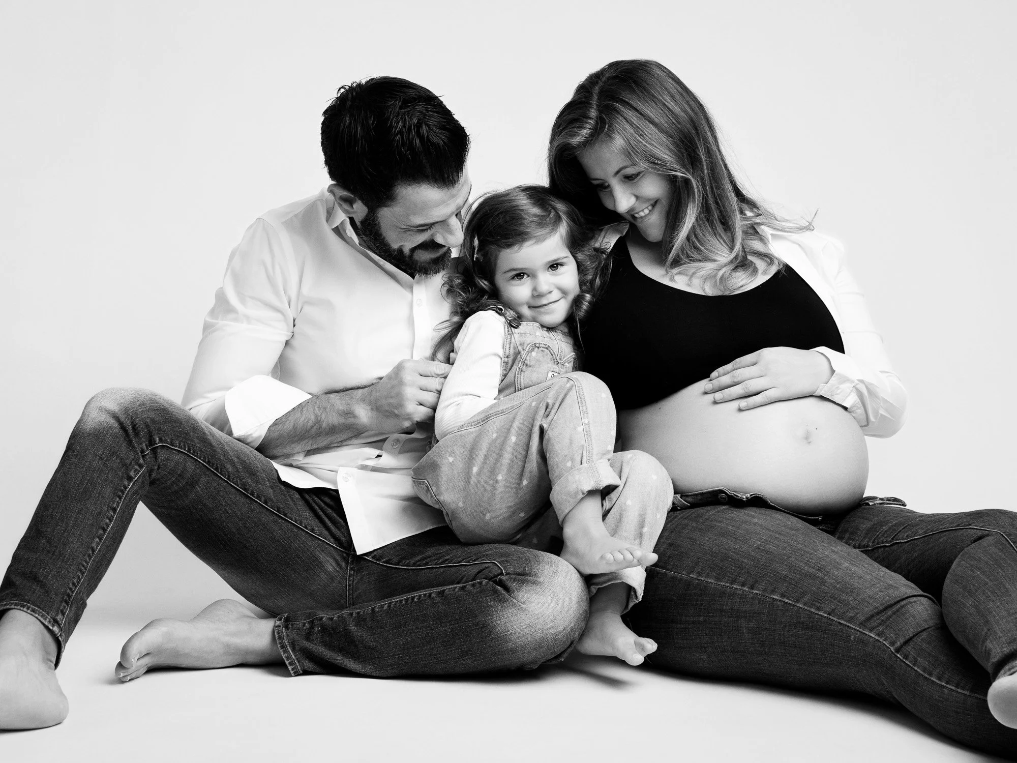 A family of three sitting together, with a pregnant woman, a man, and a young girl, all smiling and sitting on the floor.