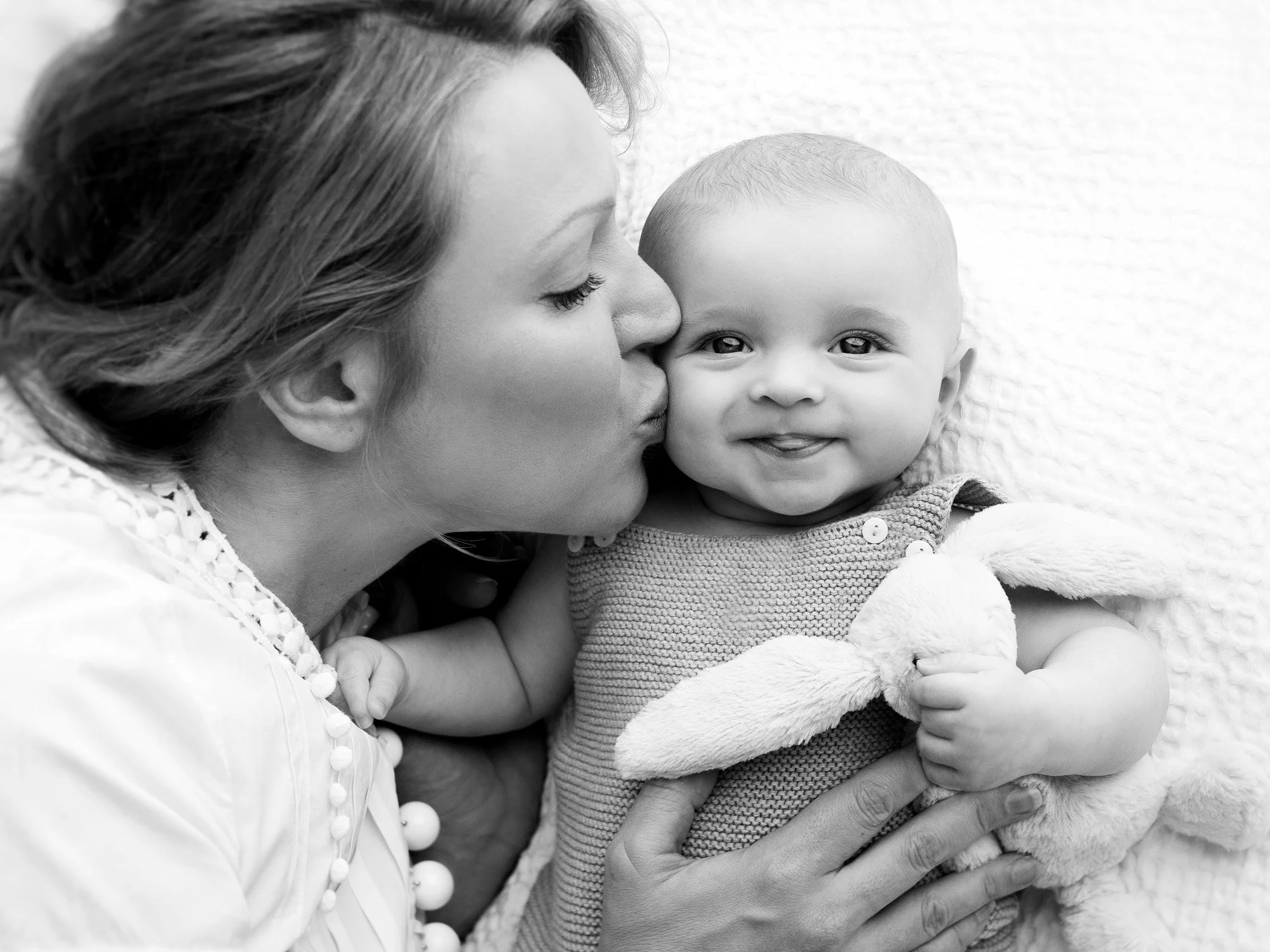 A woman is kissing a smiling baby who is holding a stuffed bunny.