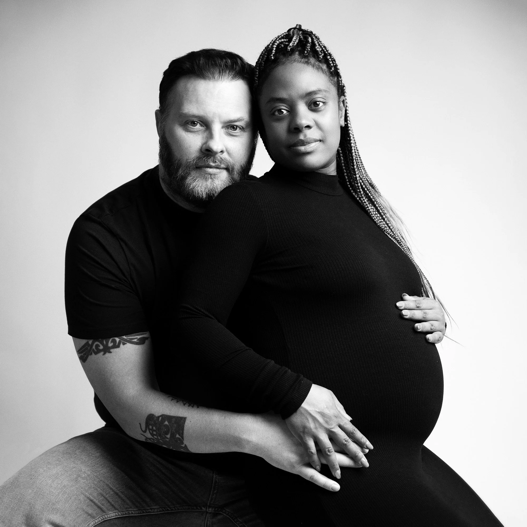 Black and white portrait of a man and pregnant woman in close pose, the man behind the woman with hands on her pregnant belly, both looking at the camera against plain light background.