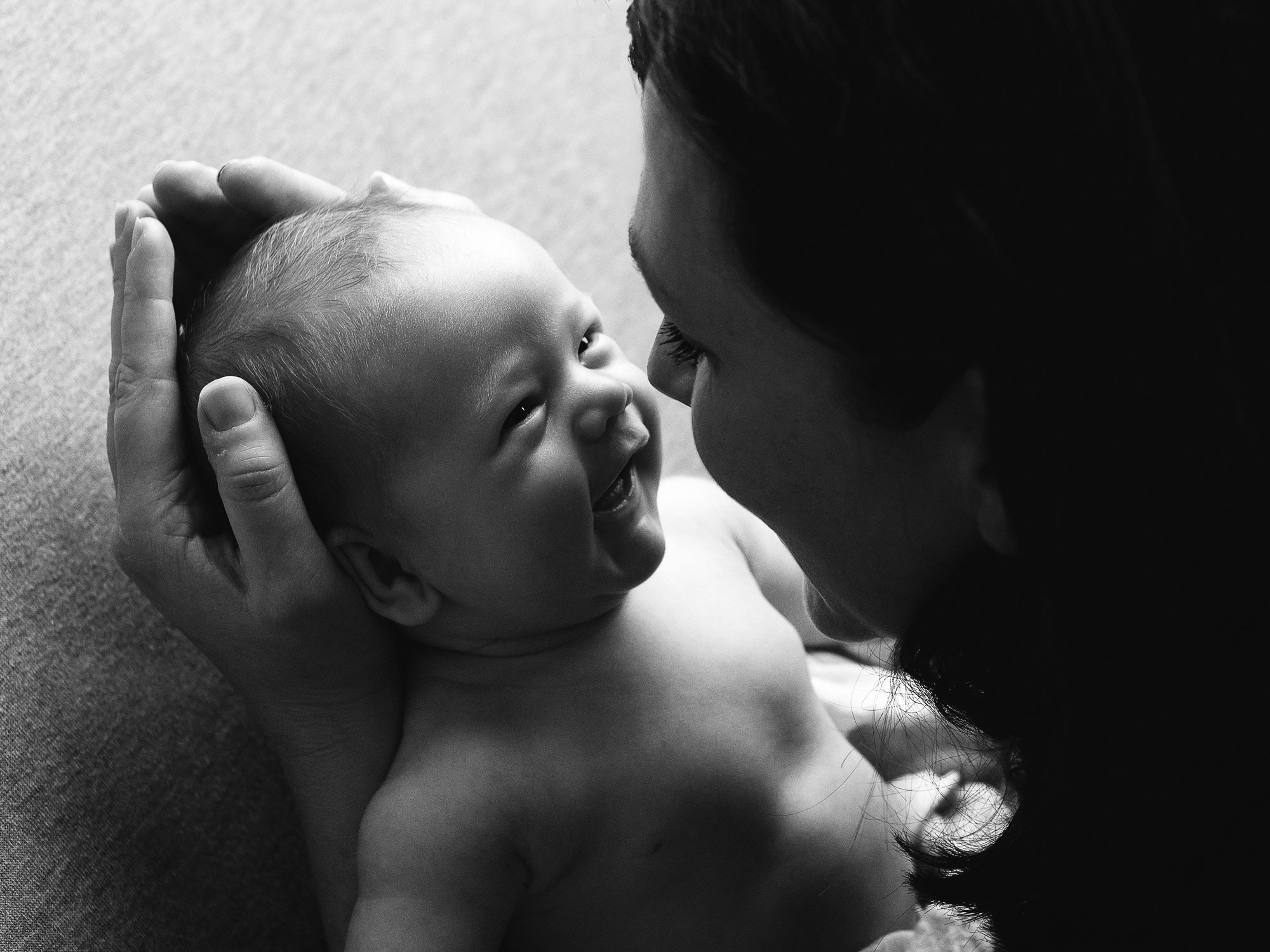 A smiling baby being held and touched gently on the head by a woman, both lying on a soft surface, with a close-up view of their faces.