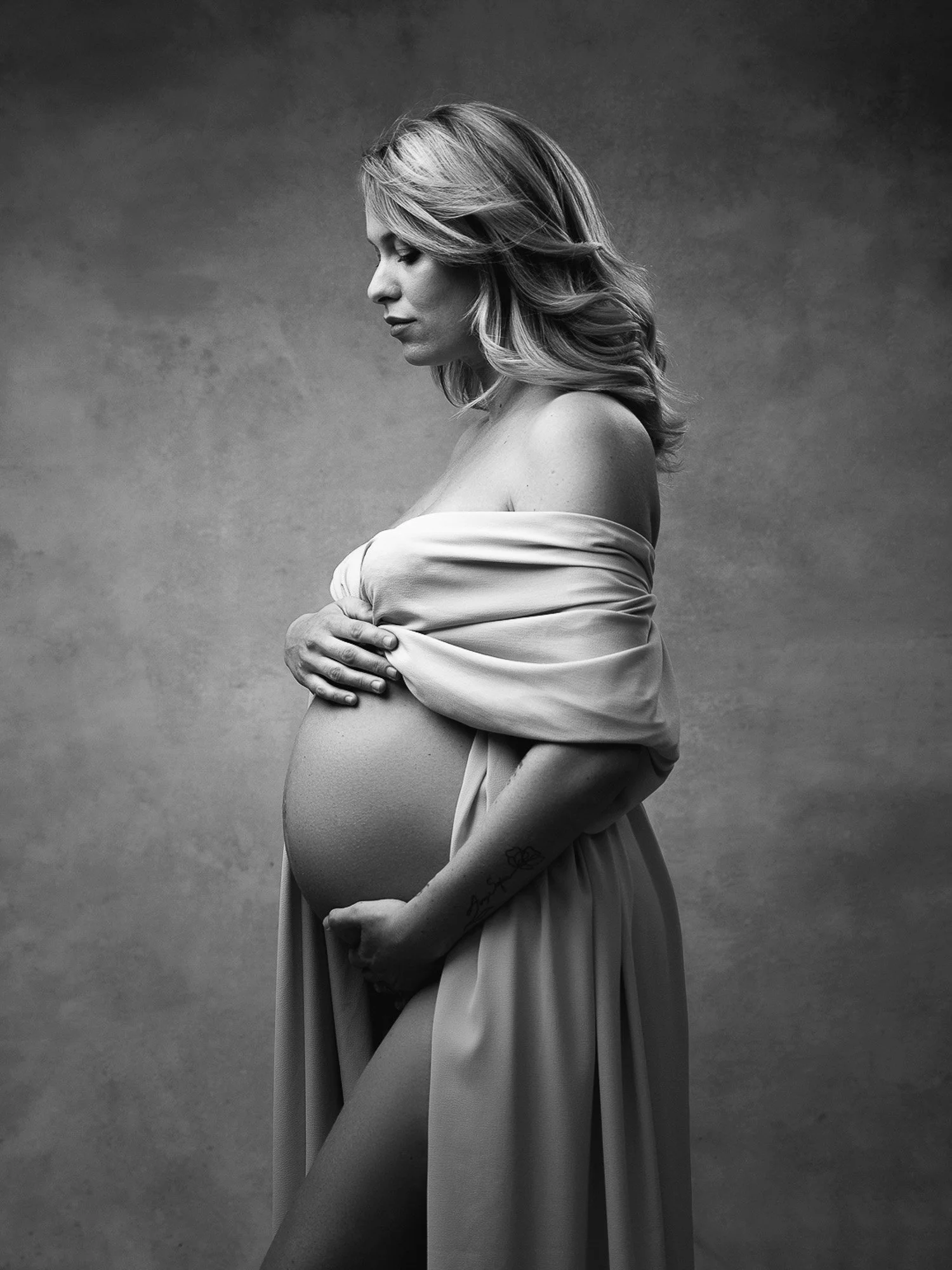 A pregnant woman with wavy blonde hair, holding her belly with one hand and covering her chest with a fabric, standing in profile against a plain background.