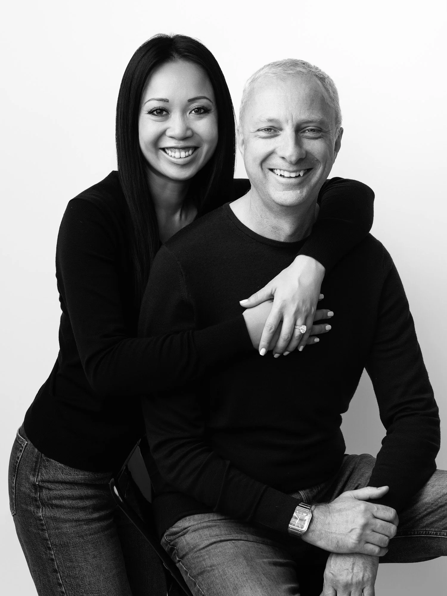A woman with long dark hair and a man with short light hair smiling, posing together in black clothing against a plain background.