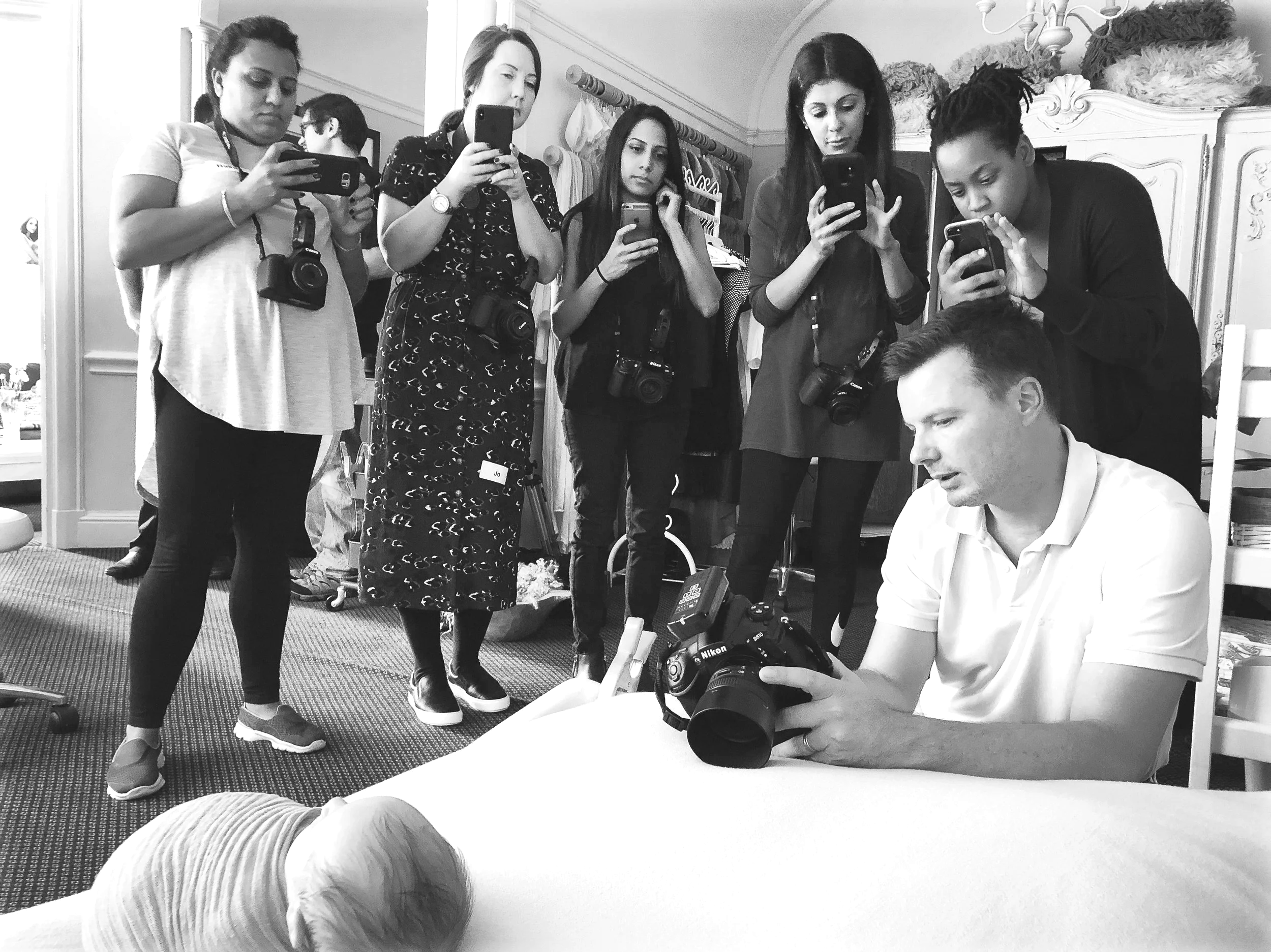 A man sitting at a table showing a camera to a group of women and men, who are all holding their phones and cameras, taking pictures or videos of a newborn baby lying on the table.