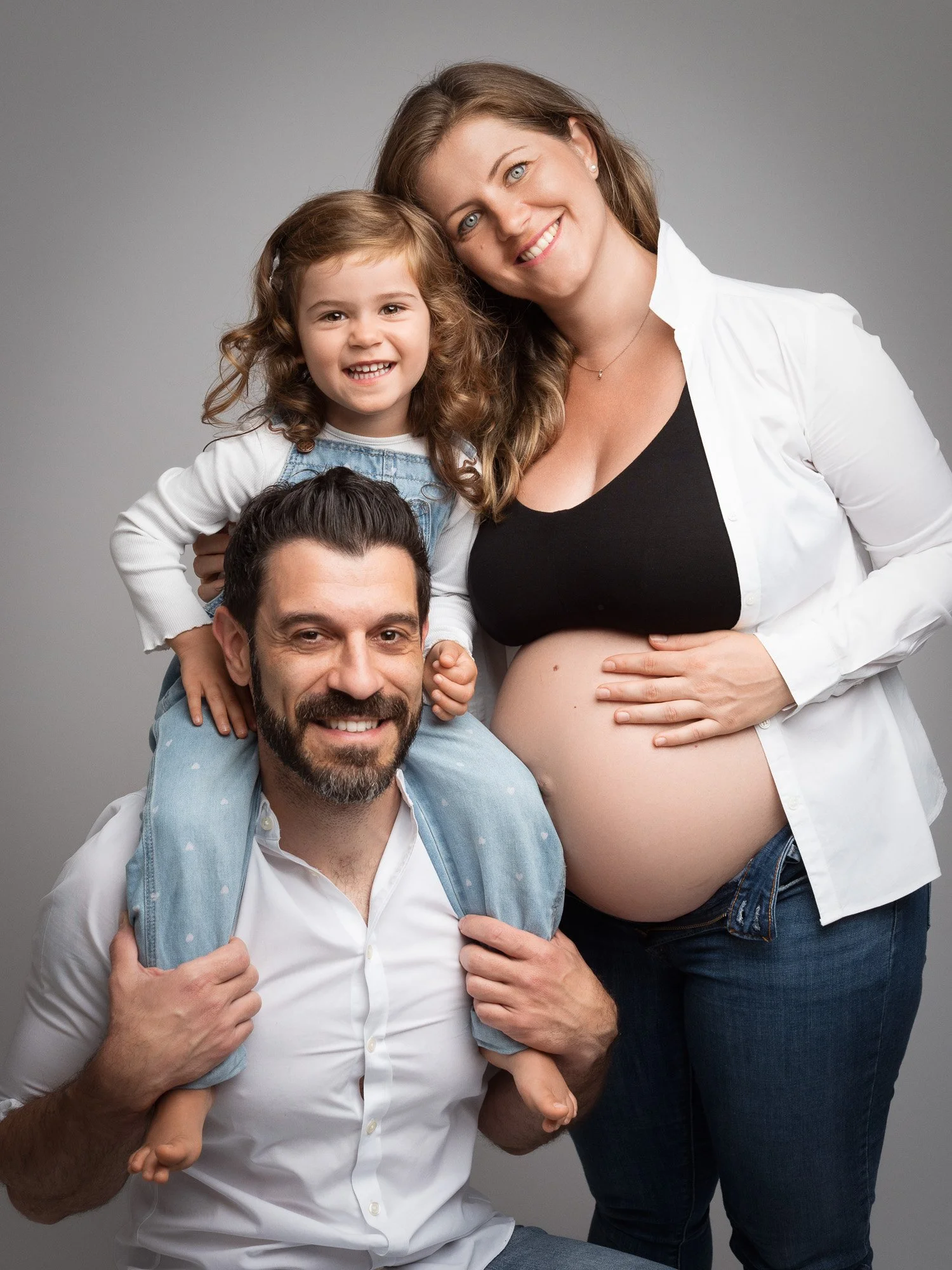 A happy family portrait featuring a pregnant woman, a man, and a young girl against a plain gray background.