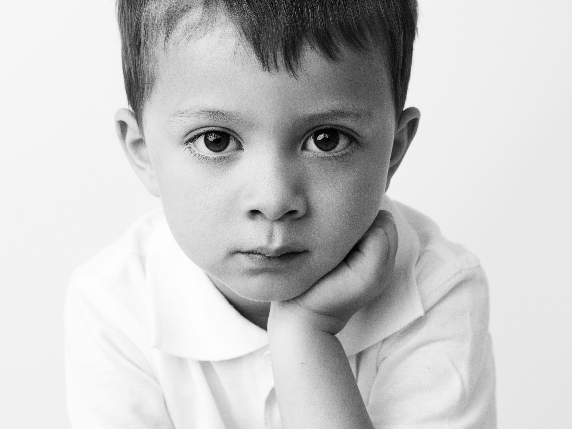 A black and white close-up portrait of a young boy with large eyes, resting his chin on his hand, wearing a collared shirt.