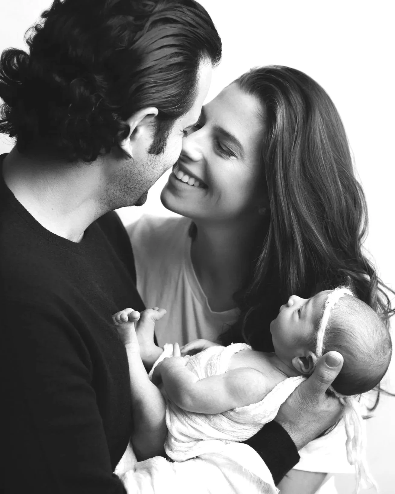 A black and white photo of a happy couple holding their sleeping baby close to their faces, smiling warmly.
