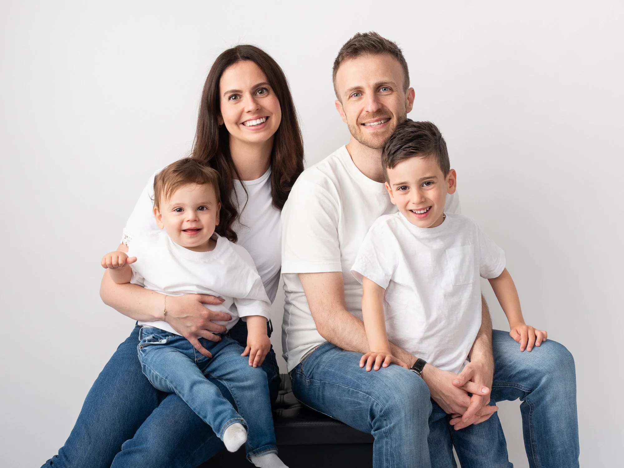 A happy family of four, including a mother, father, and two young children, sitting together against a plain white background.