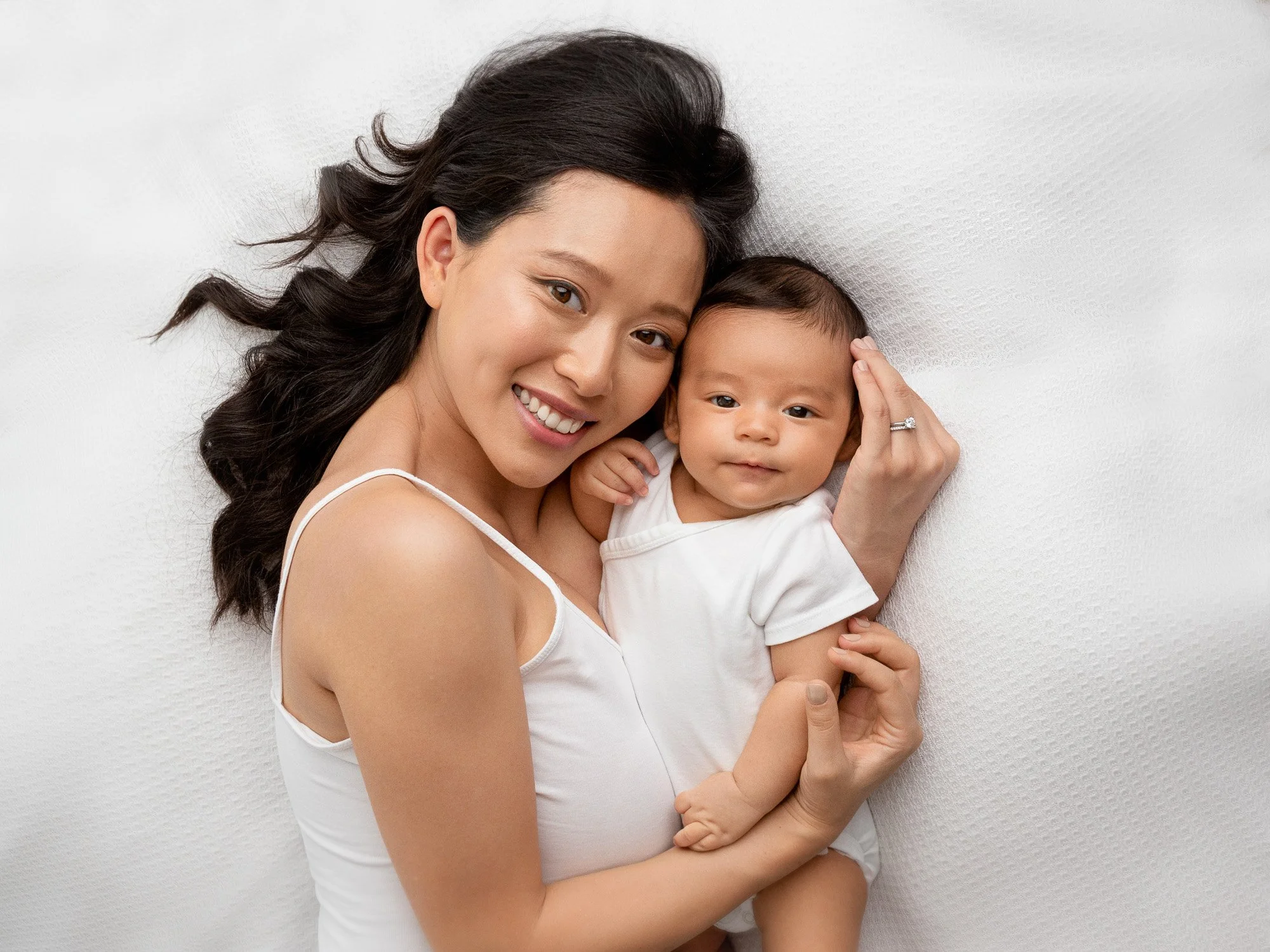 A woman with long dark hair lying on a white surface, holding a smiling baby with similar hair color, both wearing white clothing.