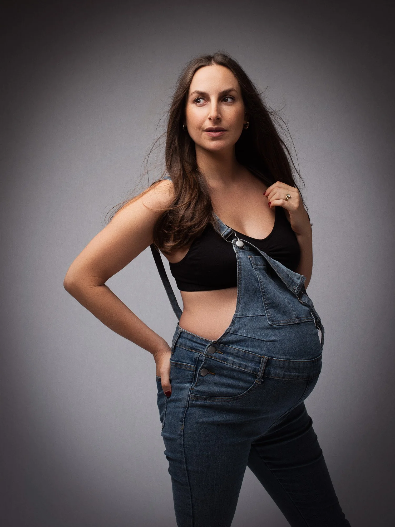 A woman with long brown hair wearing a black top and denim overalls looks to the side against a gray background.
